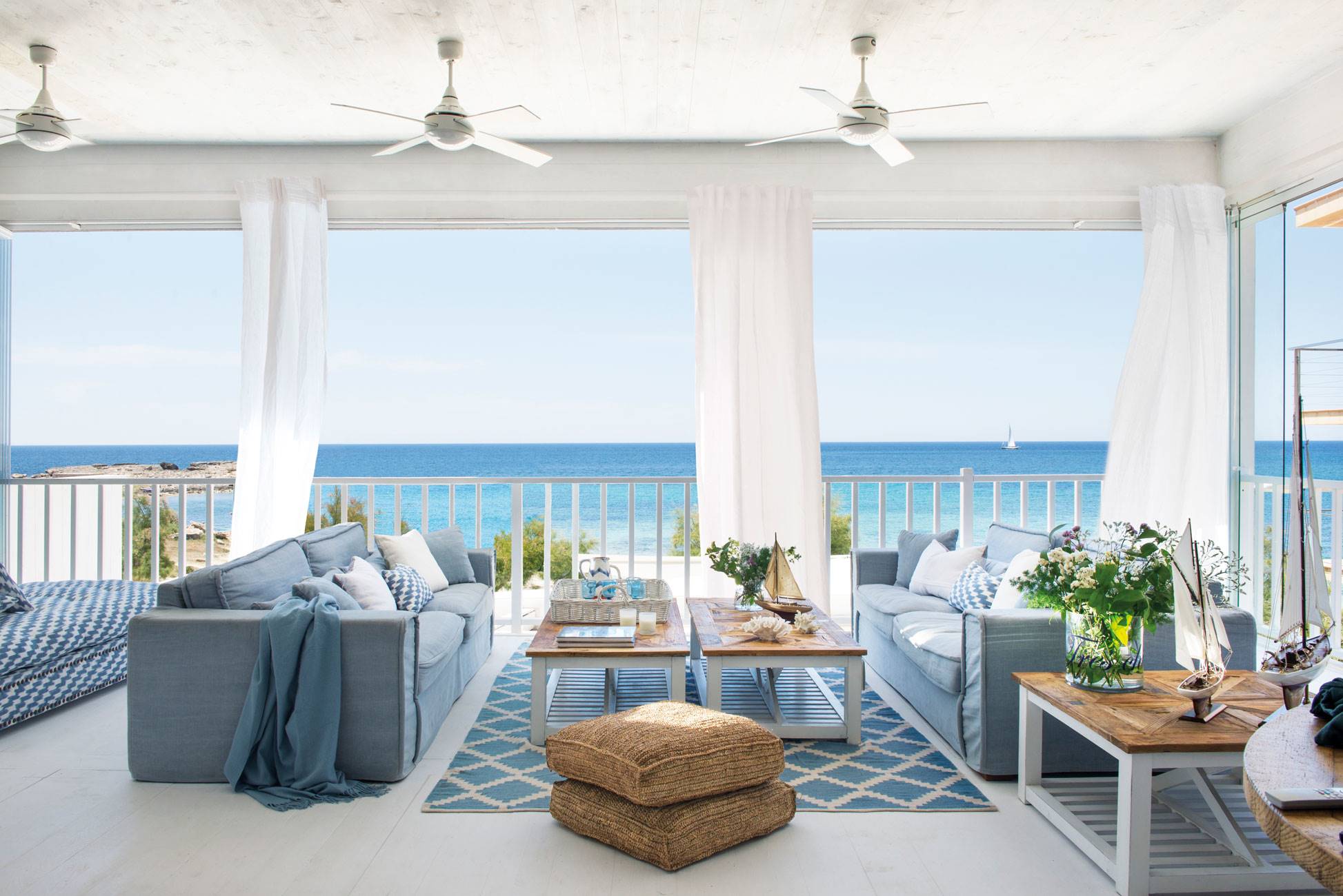 Salón de verano con vistas al mar con sofás azules y ventiladores de techo. 
