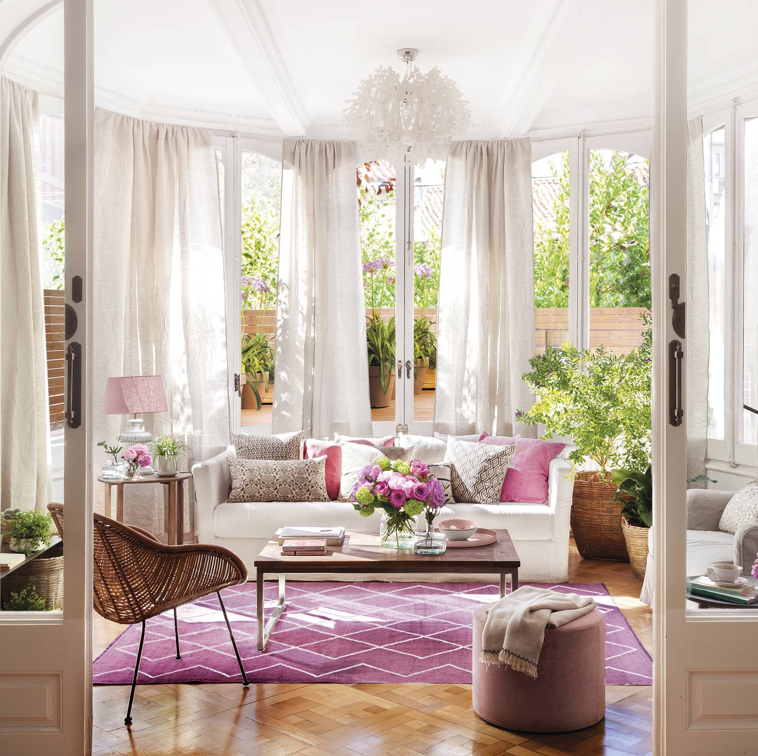 Sofá blanco con cojines y alfombra rosa en salón tipo galerí con puertas correderas con cristal, lámpara de techo, pufs y silla de fibra