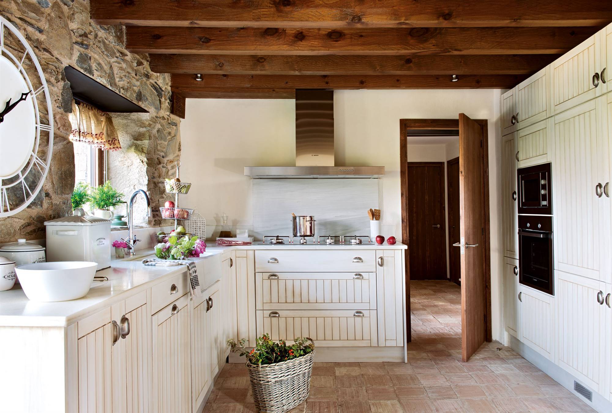 Cocina rústica con toques modernos con muebles blancos acanalados, pared de piedra y vigas de madera. 