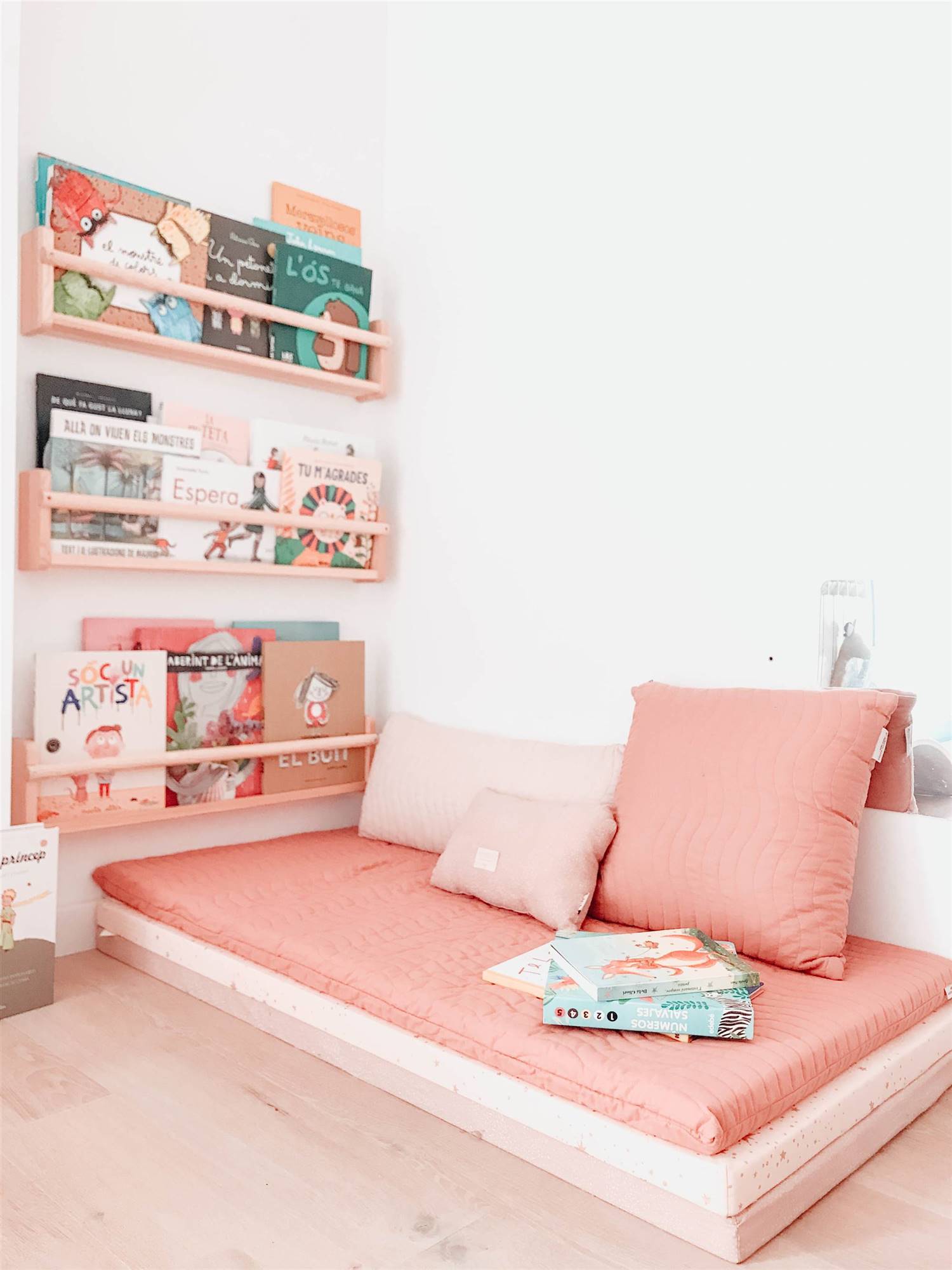 Rincón de lectura infantil decorado en color coral con un colchón, cojines y tres repisas para libros