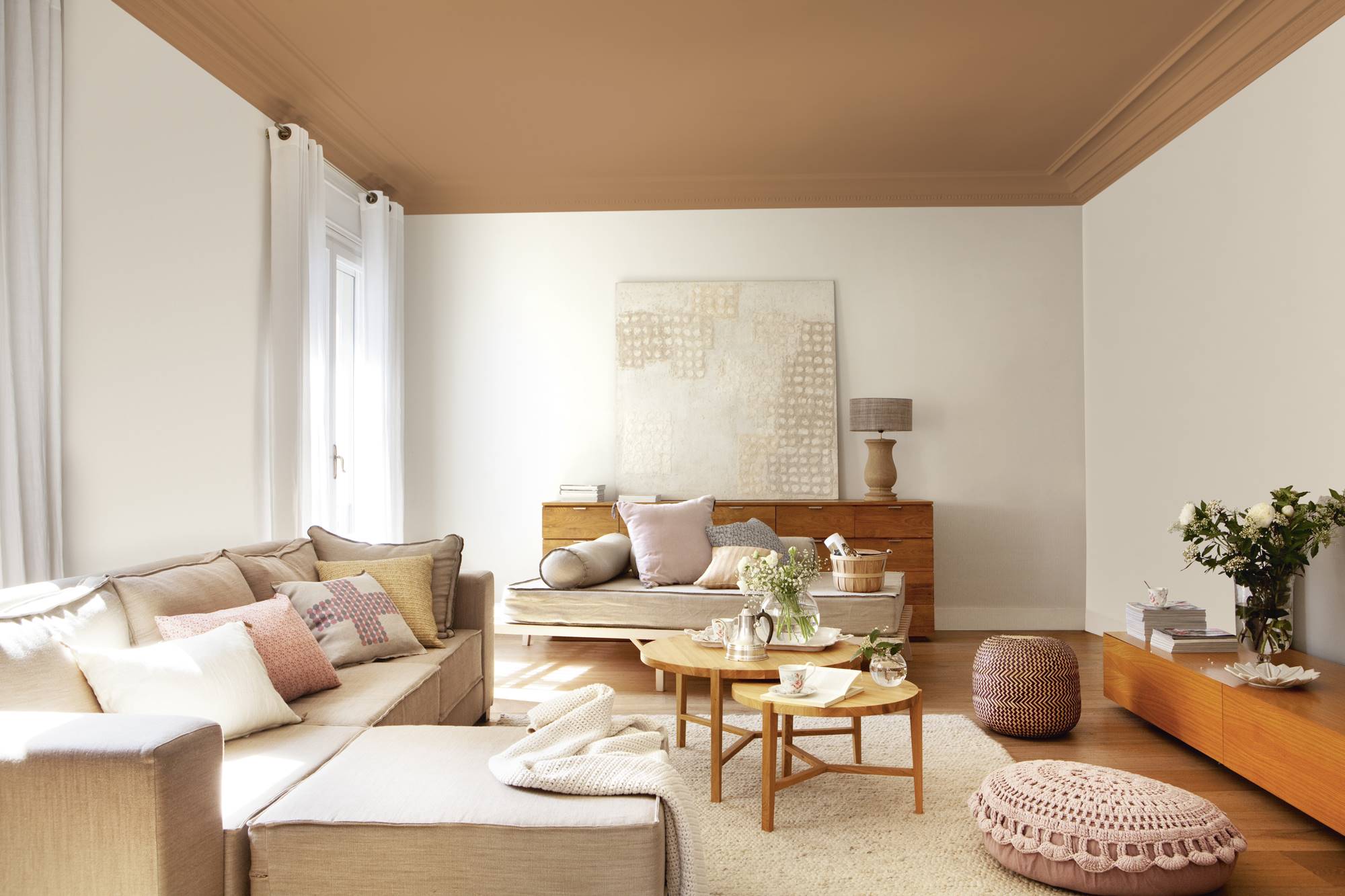 Salón con paredes blancas y techo marrón, sofá con chaise longue, mesas de centro nido redondas, pufs rosas y mueble vintage con cuadro