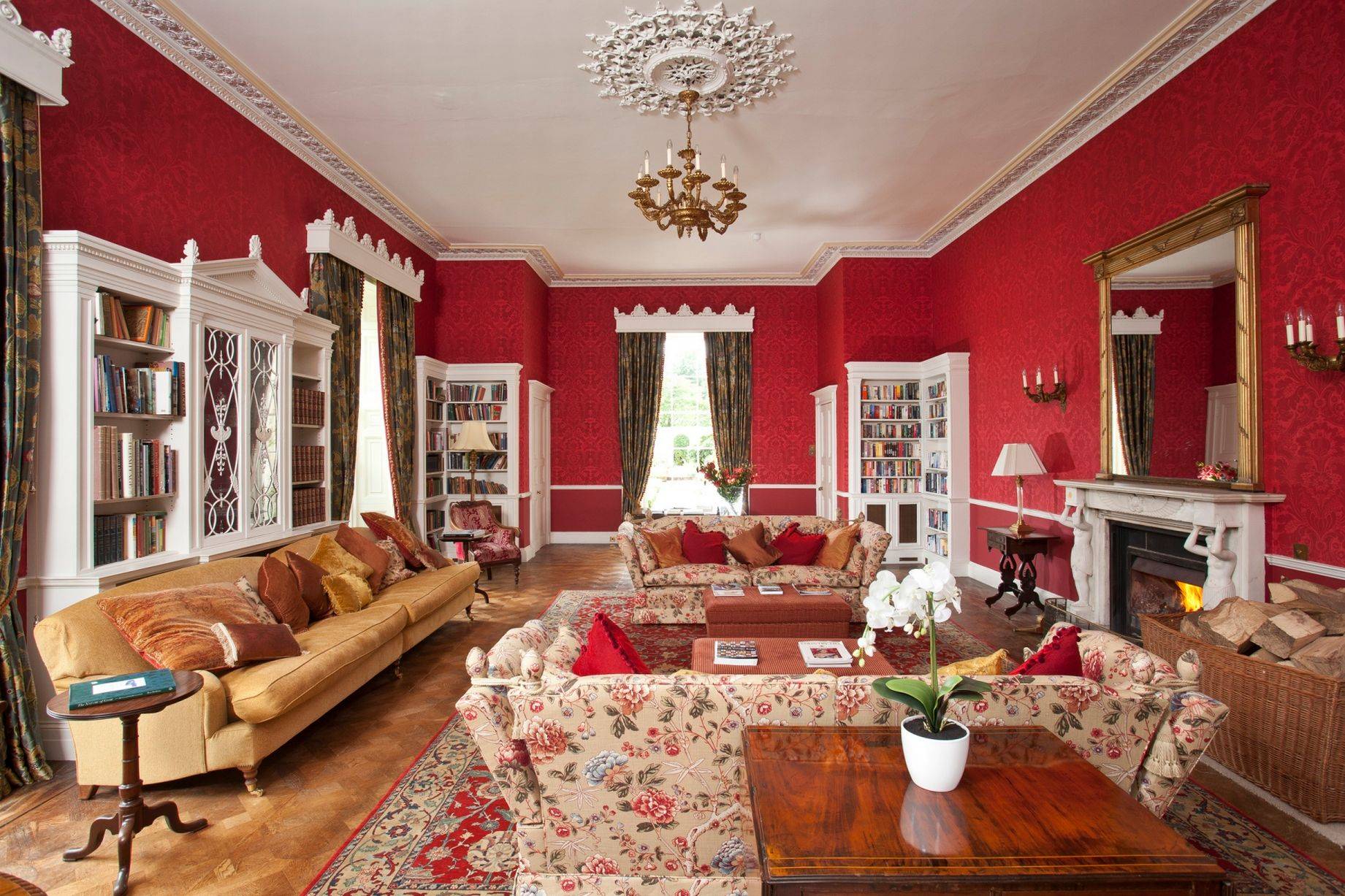 Salón con biblioteca y chimenea de piedra de estilo clásico, con papel pintado victoriano en color rojo y un rosetón en el techo