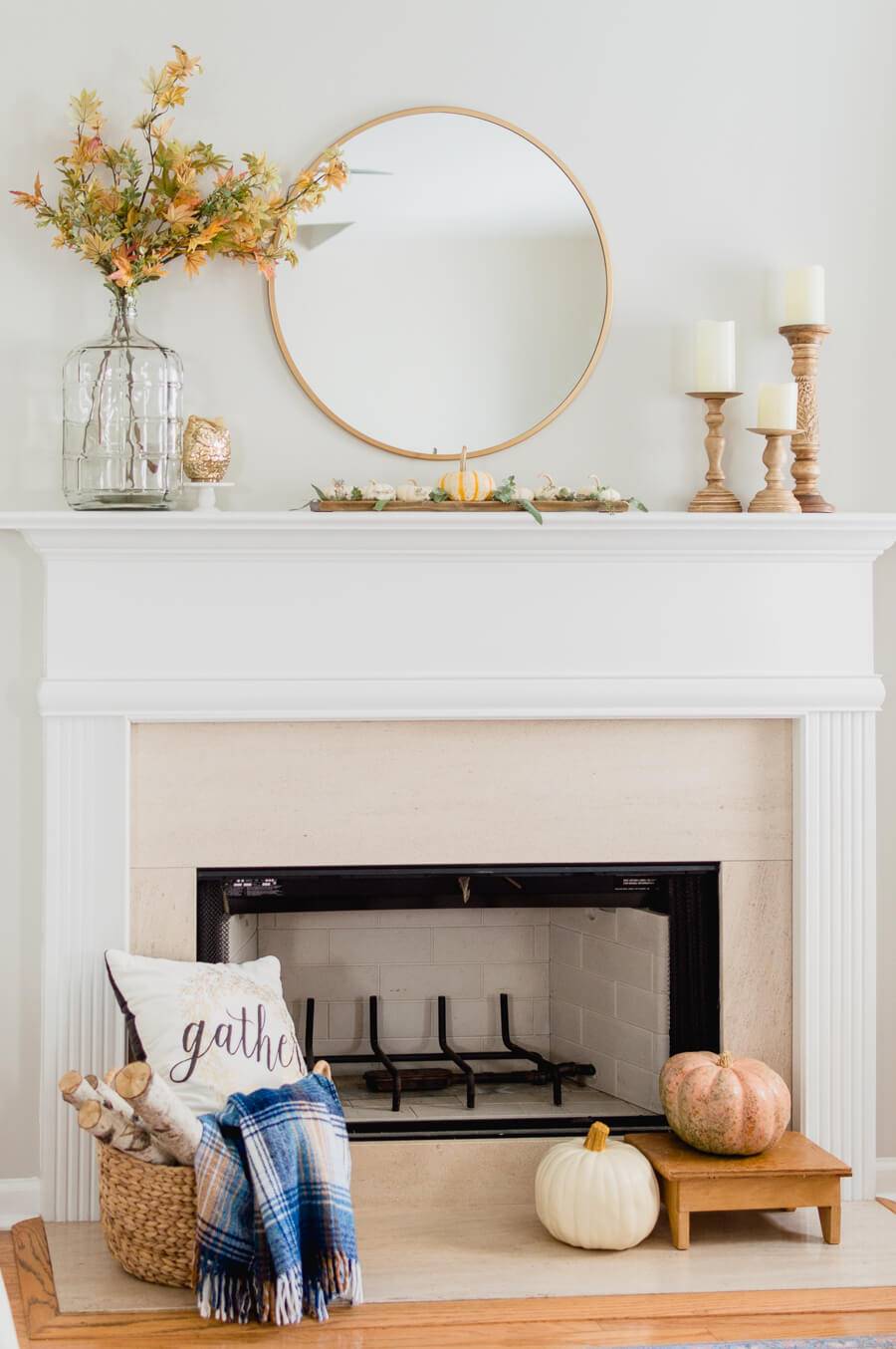 Chimenea blanca decorada con calabazas, un jarrón con hojas de otoño, un espejo redondo y portavelas de madera