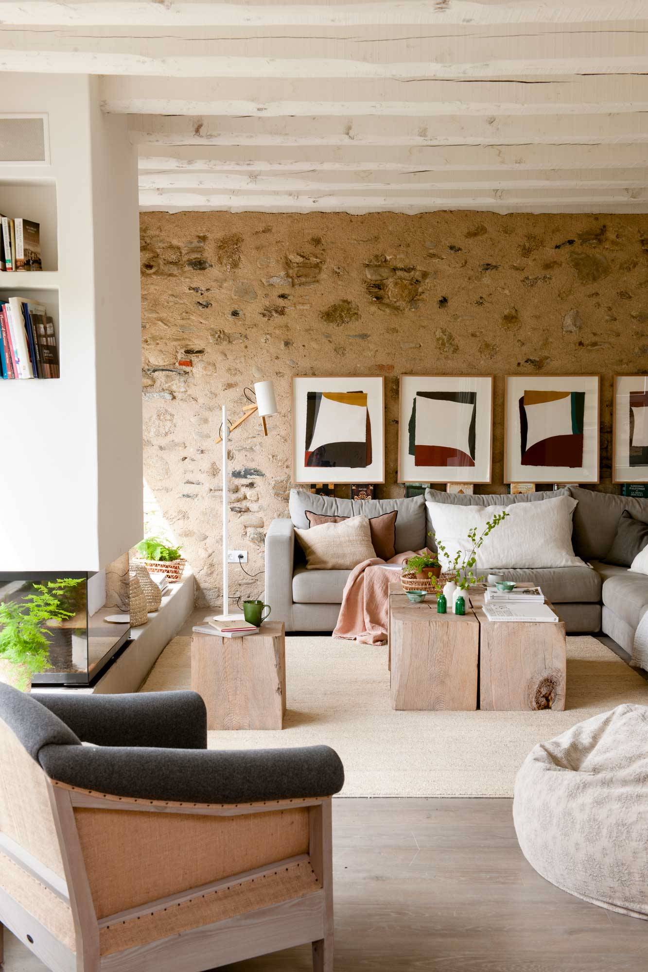 Salón con sofá gris, butaca, mesas de centro de madera rústicas y pared decorada con la piedra original y cuadros encima.