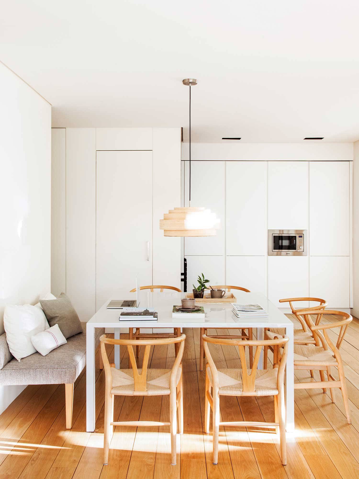 Comedor abierto a la cocina con mesa cuadrada blanca y sillas de madera. 