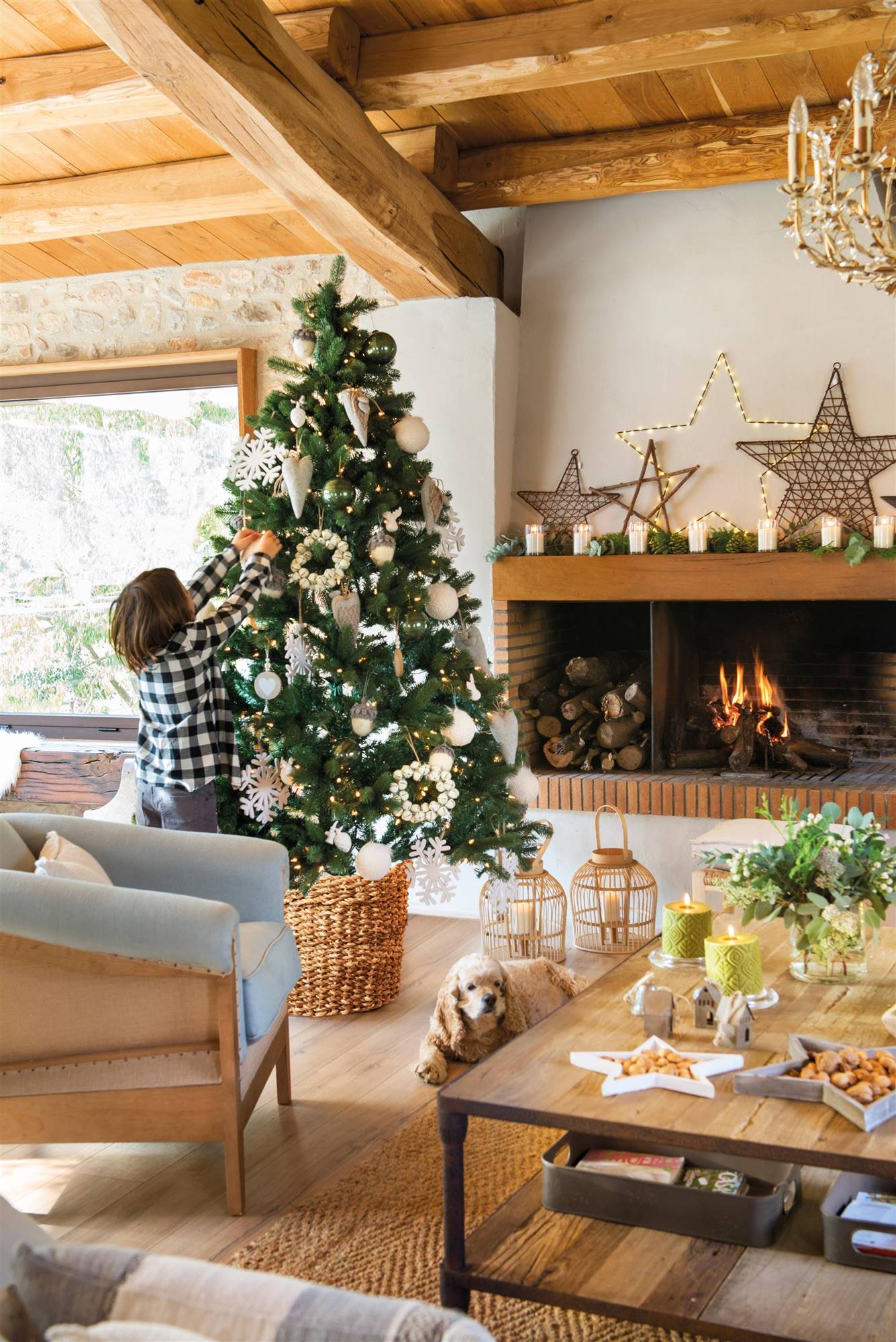 Salón rústico con árbol de Navidad decorado cerca de la chimenea y techo de vigas de madera. 