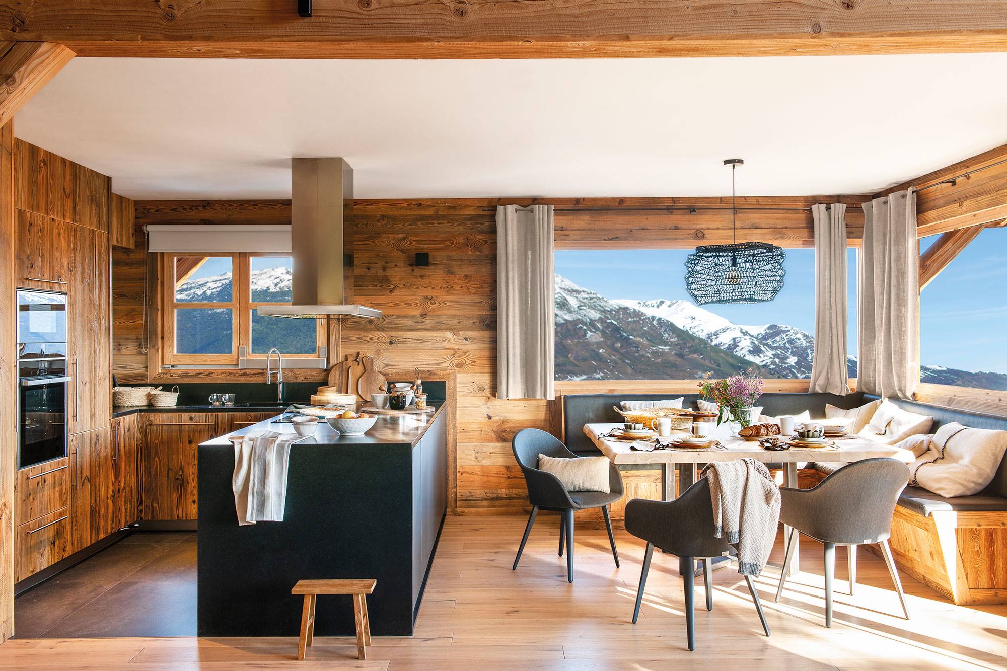 Cocina abierta al comedor con muebles negros y de madera y grandes ventanales con vistas a las montañas. 