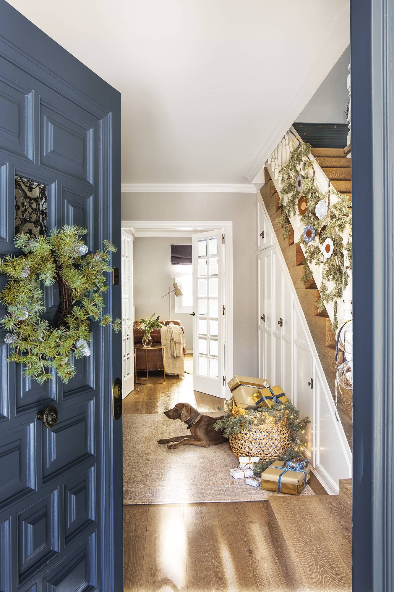 Recibidor con puerta azul con corona, guirnalda en la escalera y regalos en una cesta.