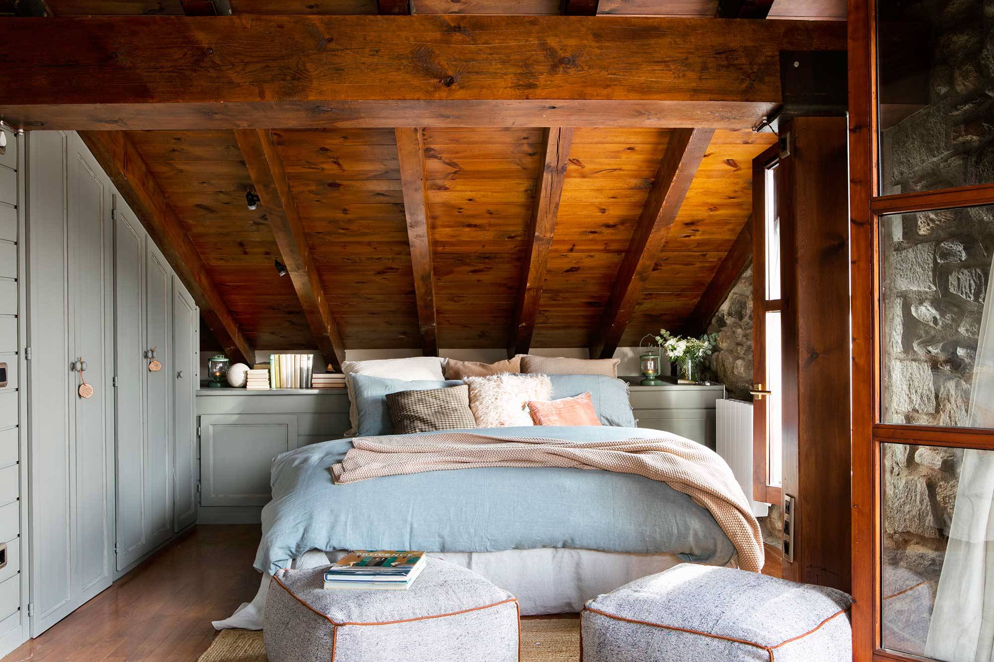 Dormitorio en buhardilla rústica revestida de madera y con puffs.