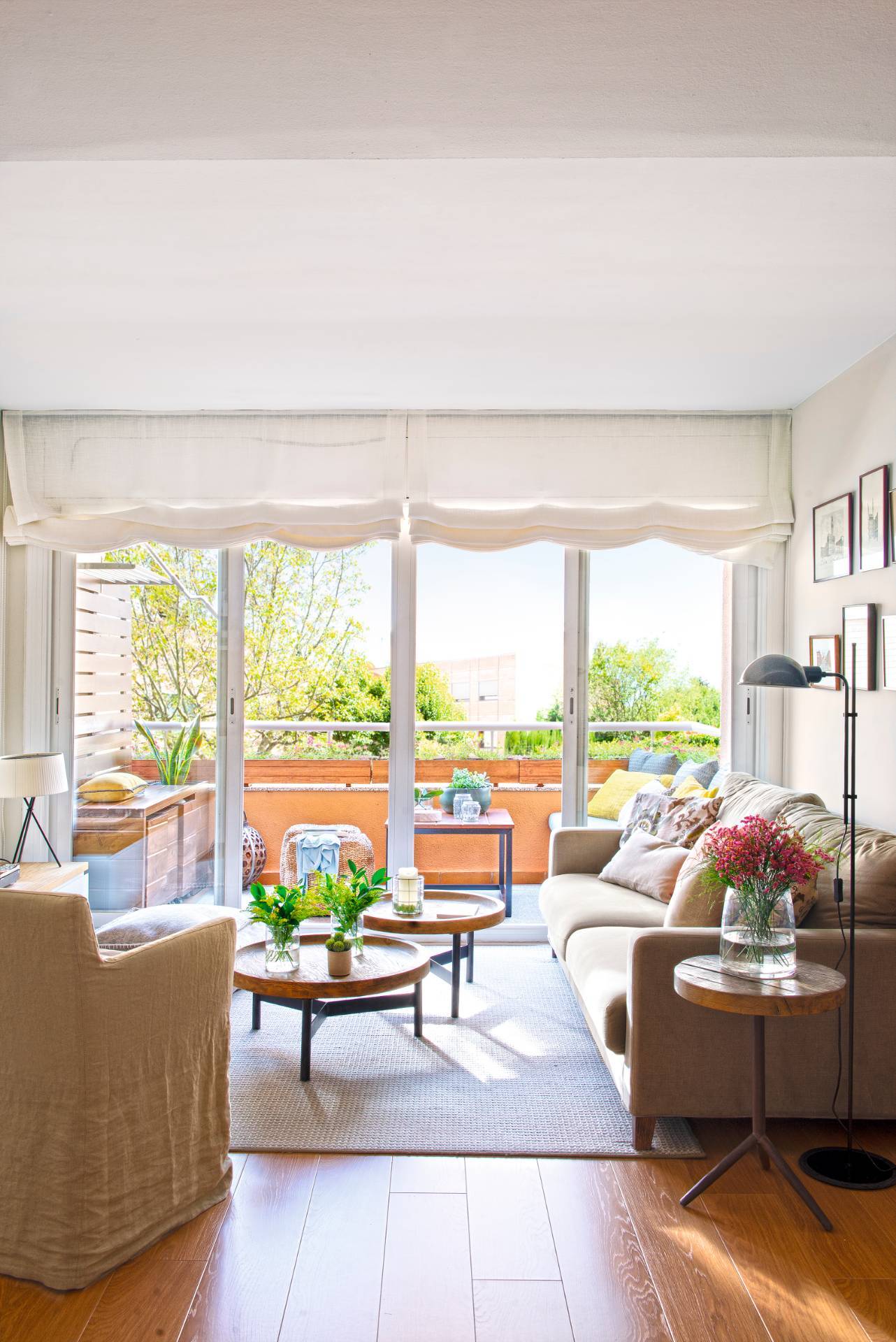 Salón pequeño con gran ventanal que da a una terraza, sofá beige y mesas de centro redondas. 
