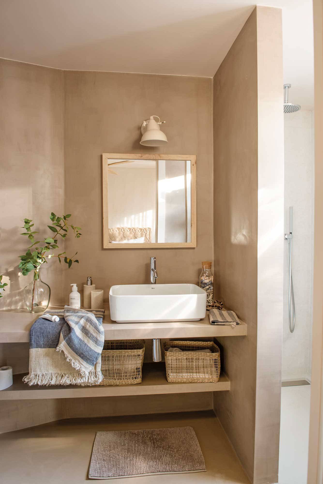Baño pequeño en tonos neutros con un armario bajolavabo, lámpara de aplique, revestimiento de microcemento y cestas de fibras naturales. 