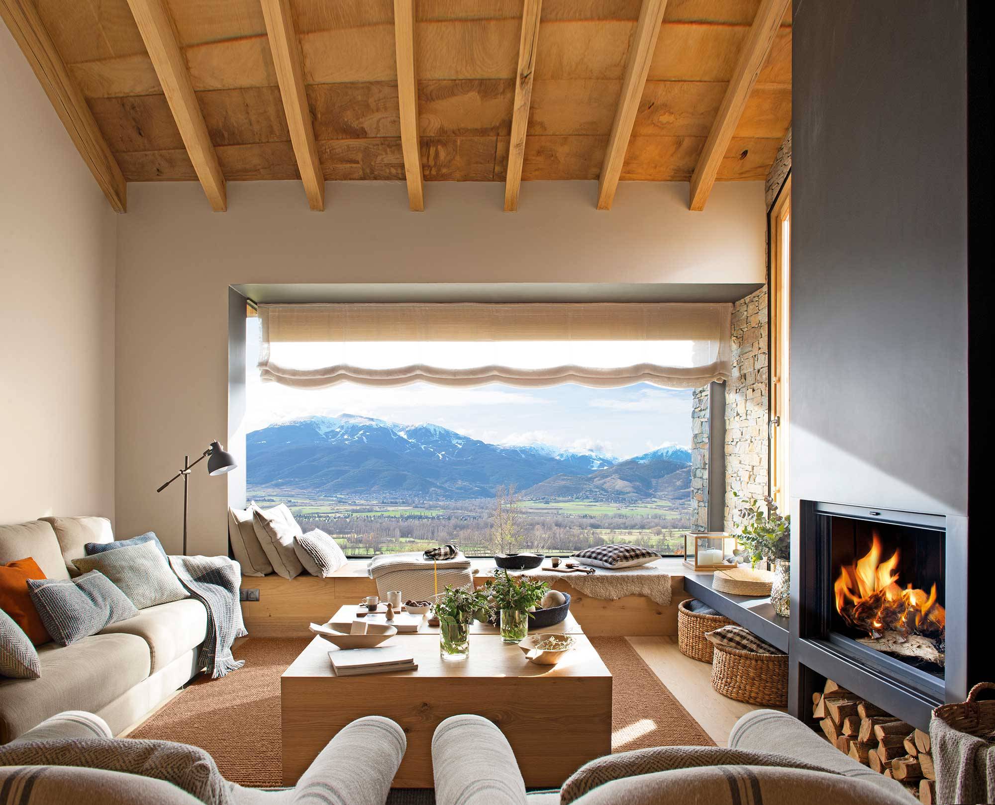 Salón de casa de montaña con sofás beige y gran ventanal.