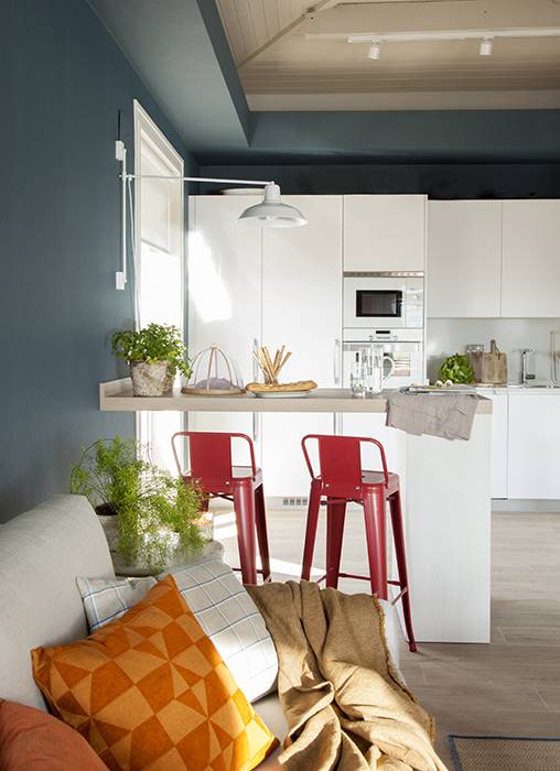 Cocina abierta con barra, taburetes rojos y paredes azules.