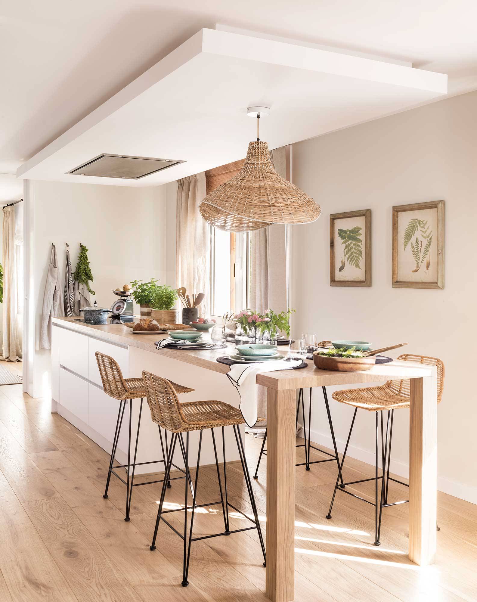 Cocina blanca moderna con isla central, mesa de comedor de madera integrada, taburetes y lámpara de fibras. 