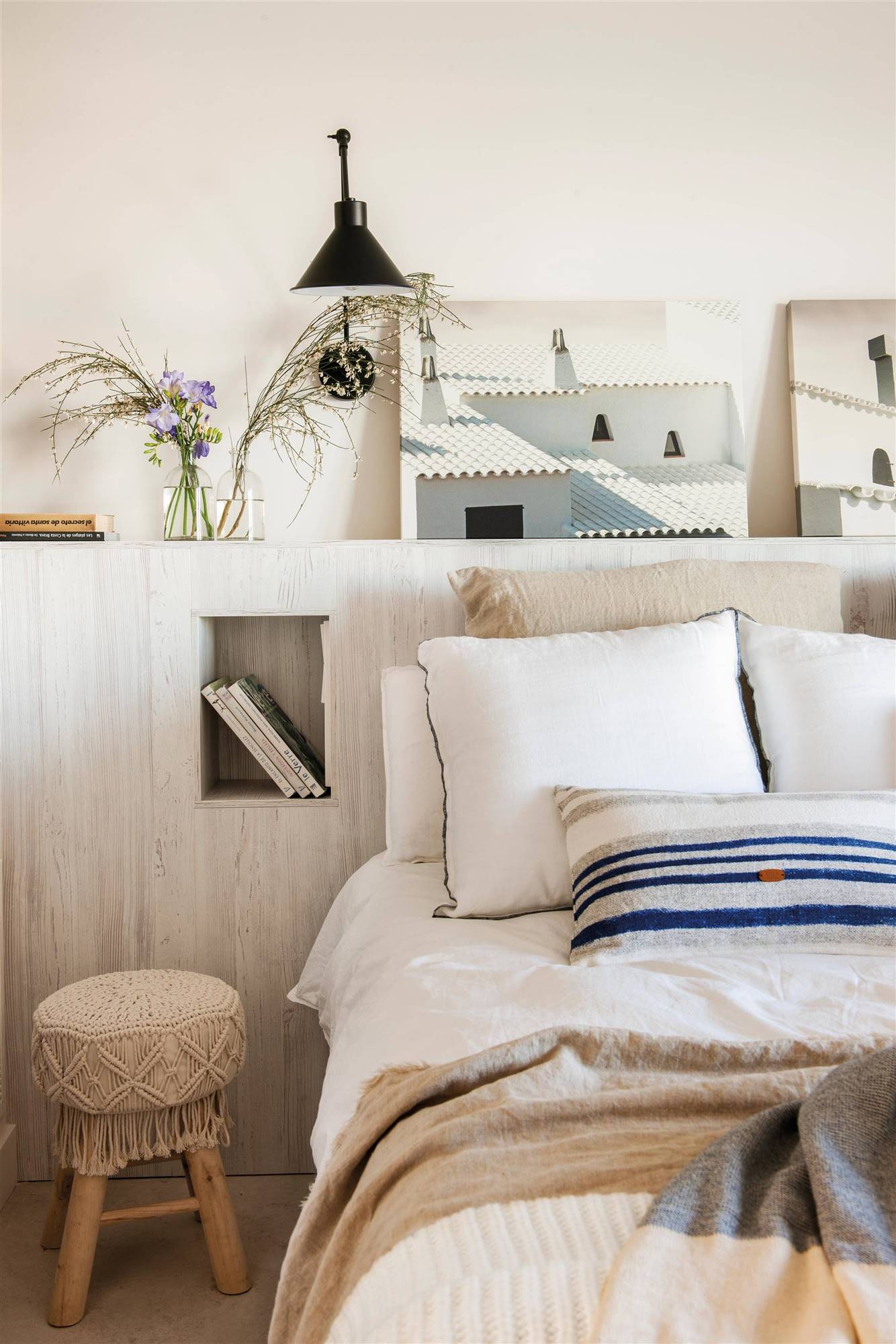 Dormitorio de primavera con cabecero de obra con mesillas integradas, taburetes de croché y cuadros de casas marineras.