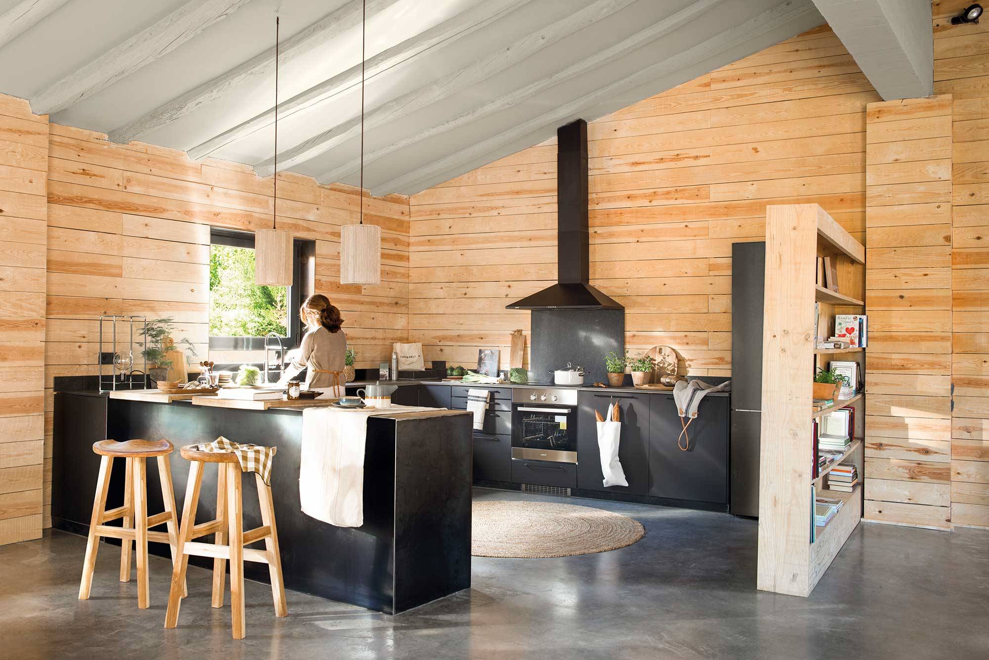 Moderna cocina con mobiliario negro, paredes de madera y taburetes de estilo rústico. 