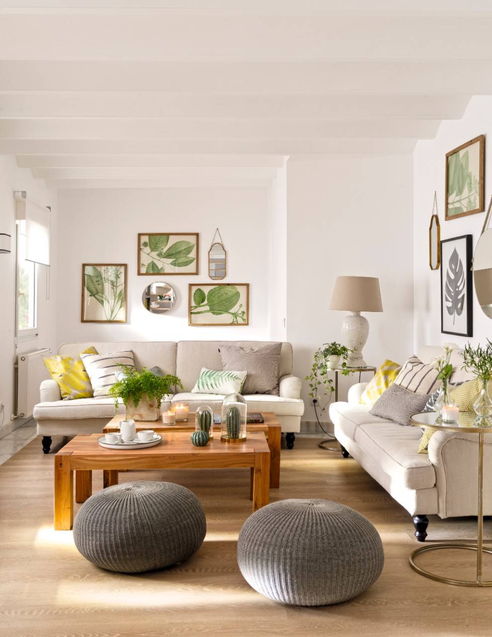 Salón con sofás y pufs grises, cuadros botánicos y cojines verdes y amarillos.