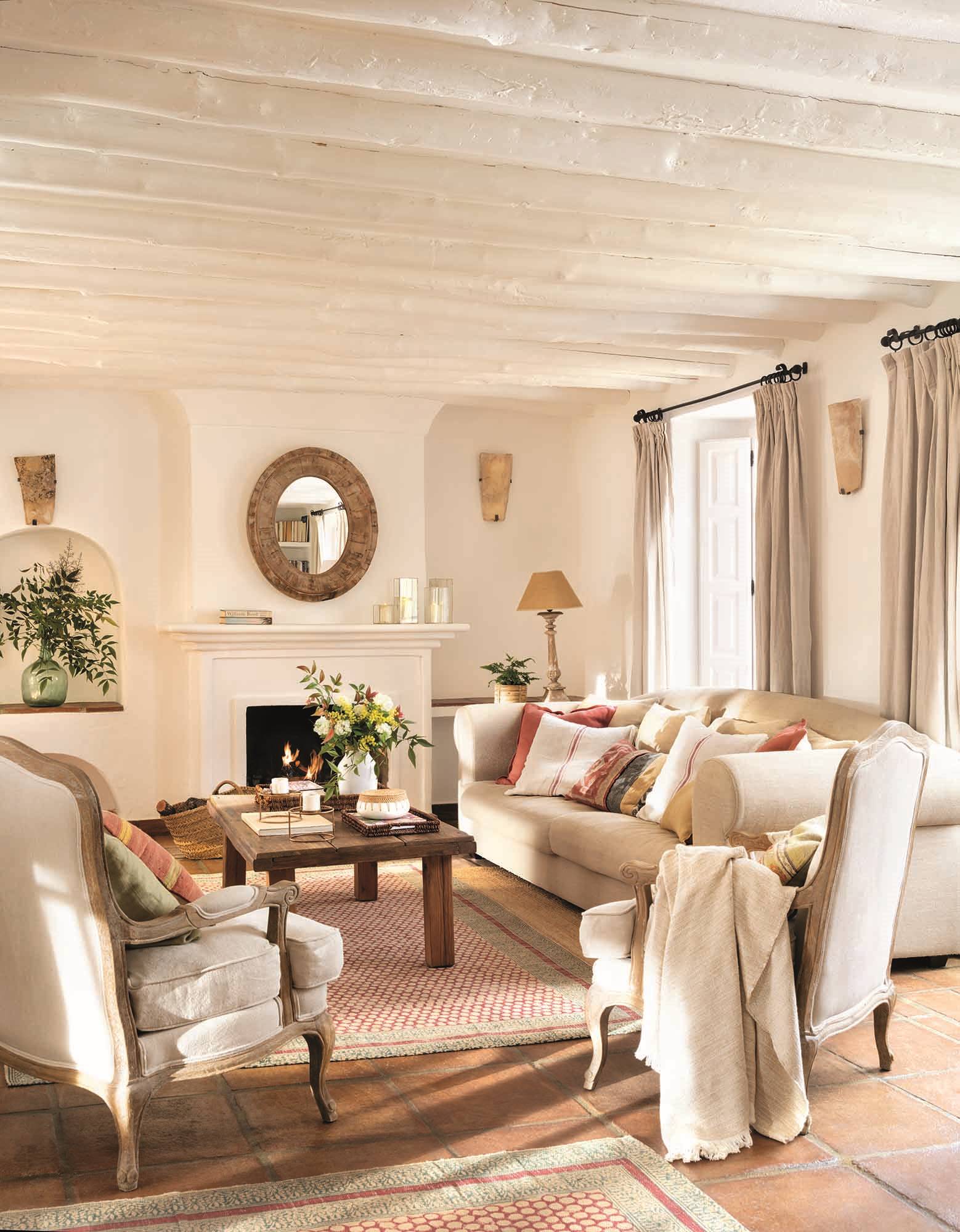 Un salón blanco con alfombras y techos con vigas, toques de color en rojo y con la madera. 