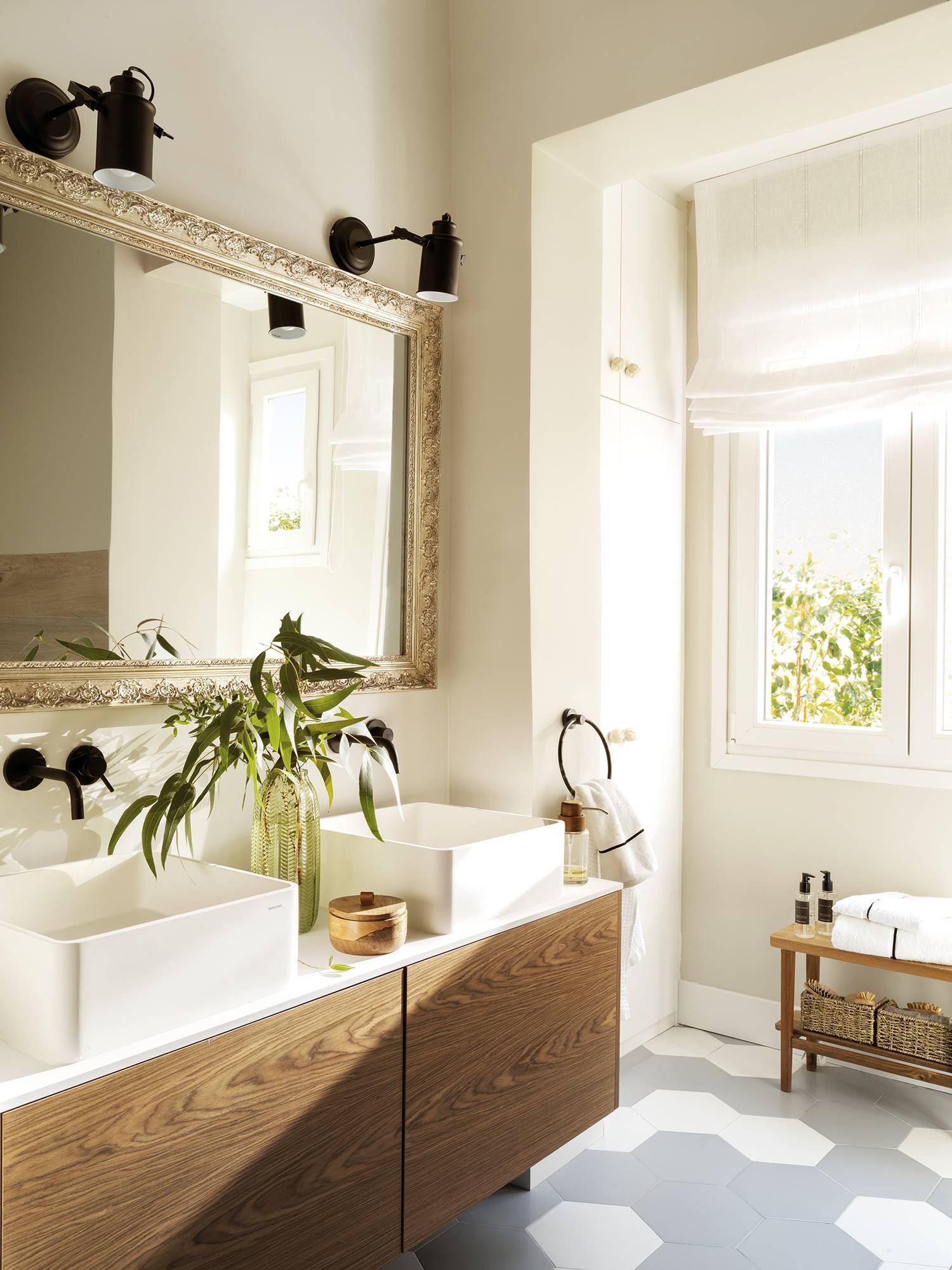 Baño con suelo de baldosas hexagonales, mueble de madera, dos lavabos y un espejo con marco dorado antiguo bajo dos apliques modernos de metal negro.