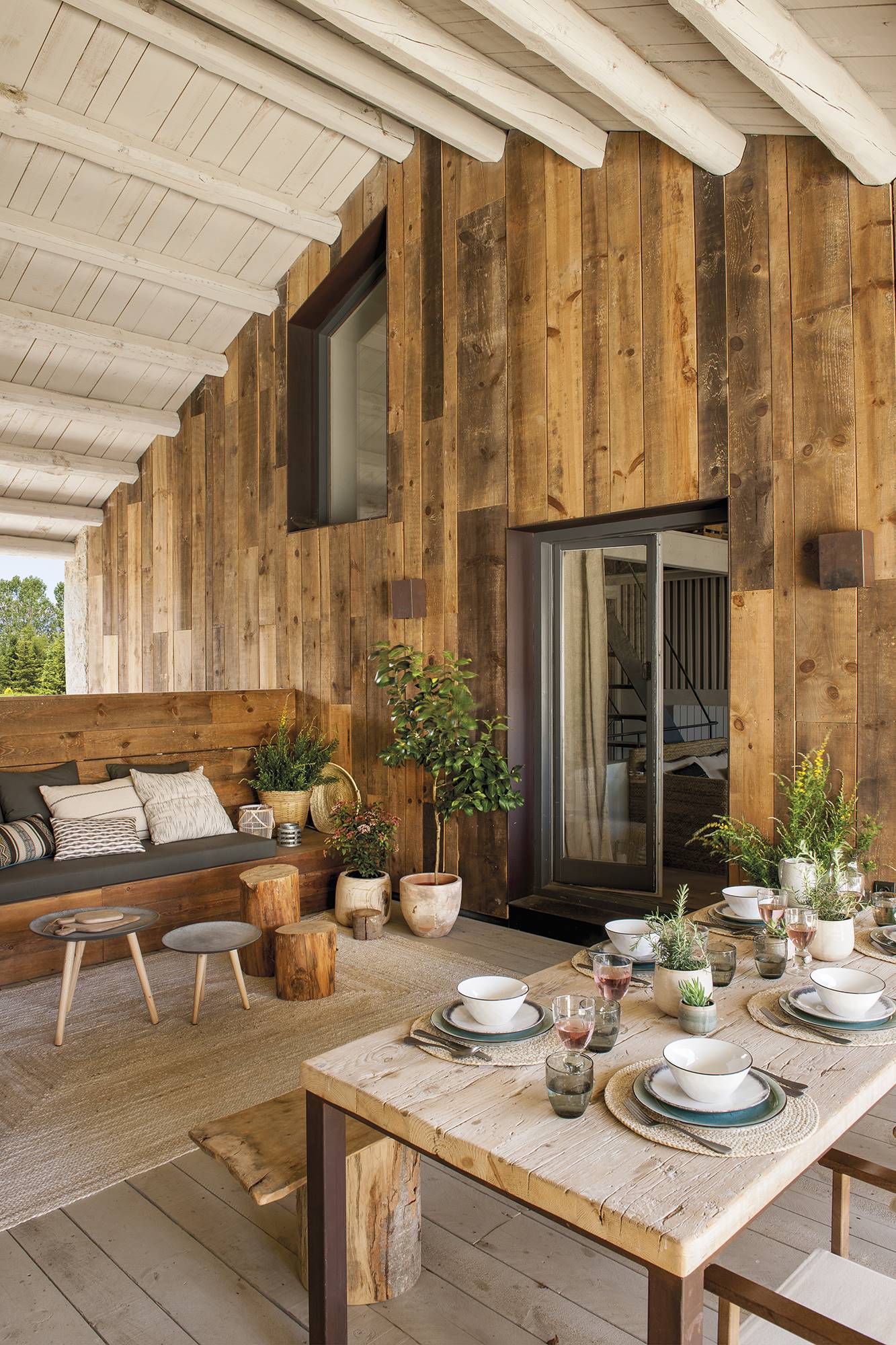 Porche de casa de montaña con paredes revestidas de madera, banco de zona de relax y comedor. 