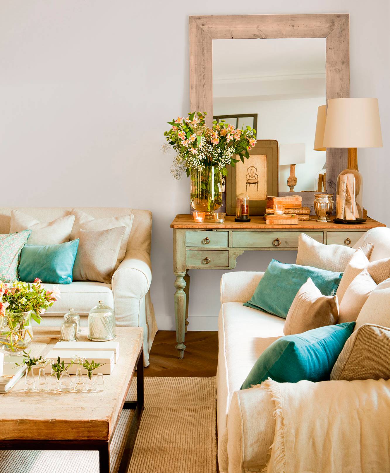 Salón con sofás beige y cojines en verde y azul, mueble decapado en verde agua, espejo y mesa de centro con detalles en cristal