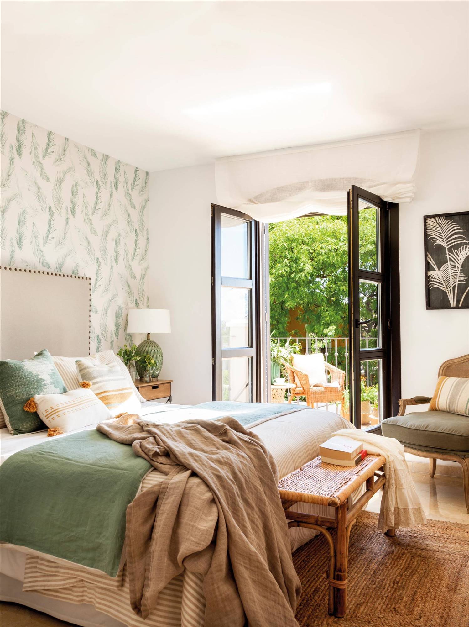 Dormitorio con cabecero tapizado con tachuelas,, banco de fibra y papel pintado con motivos vegetales en la pared del cabecero.  