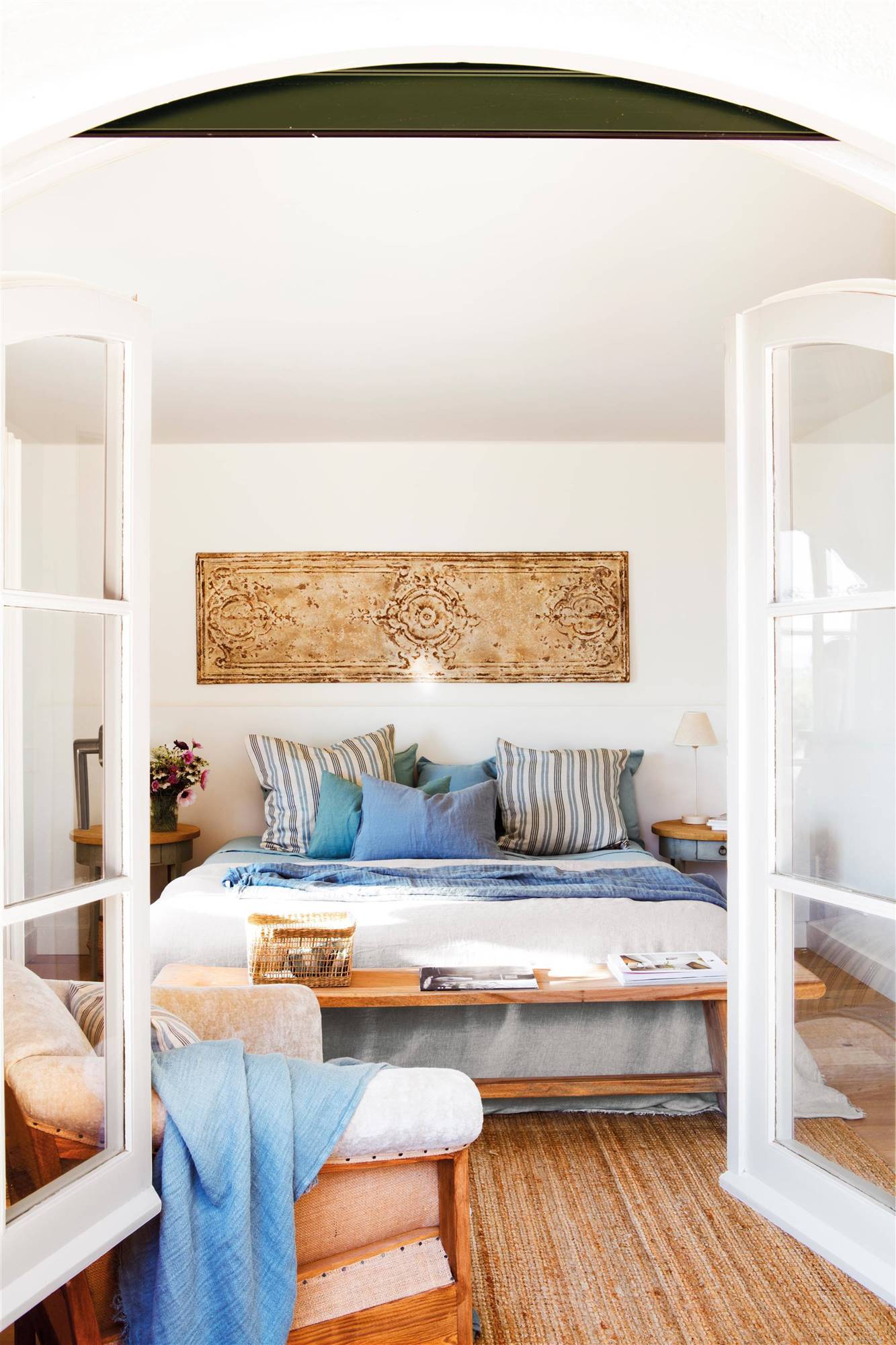 Dormitorio en blanco con ropa de cama azul y blanca y banco al pie de la cama. 
