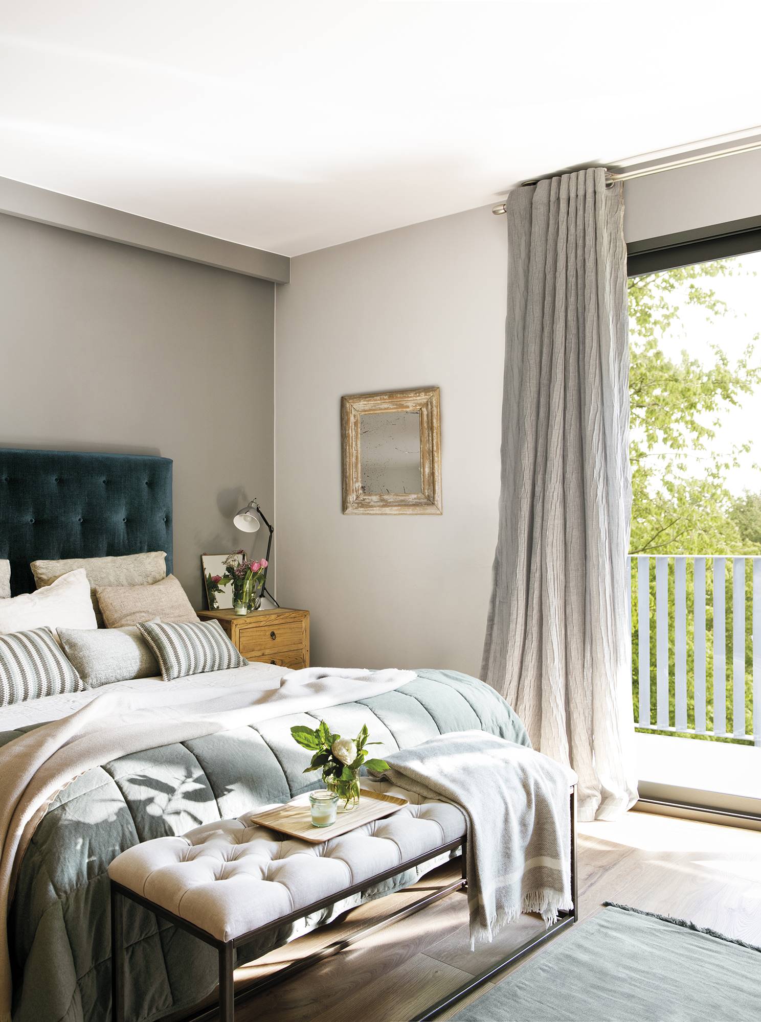 Dormitorio con cabecero tapizado, cortinas grises y banco tapizado al pie de la cama. 