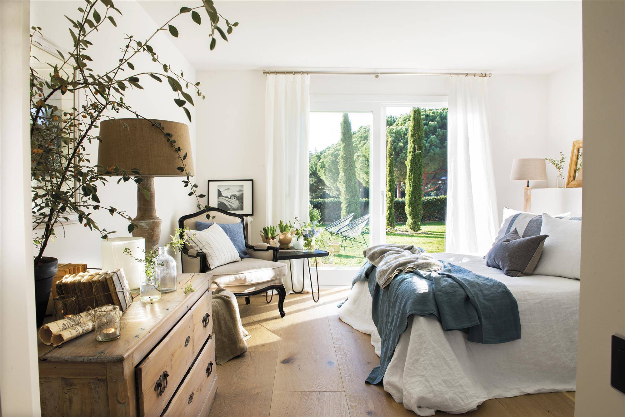 Dormitorio de verano clásico con cómoda de madera vintage, ropa de cama de lino en color blanco y azul, butaca tipo poltrona clásica y ventanas con acceso al jardín DSC2141