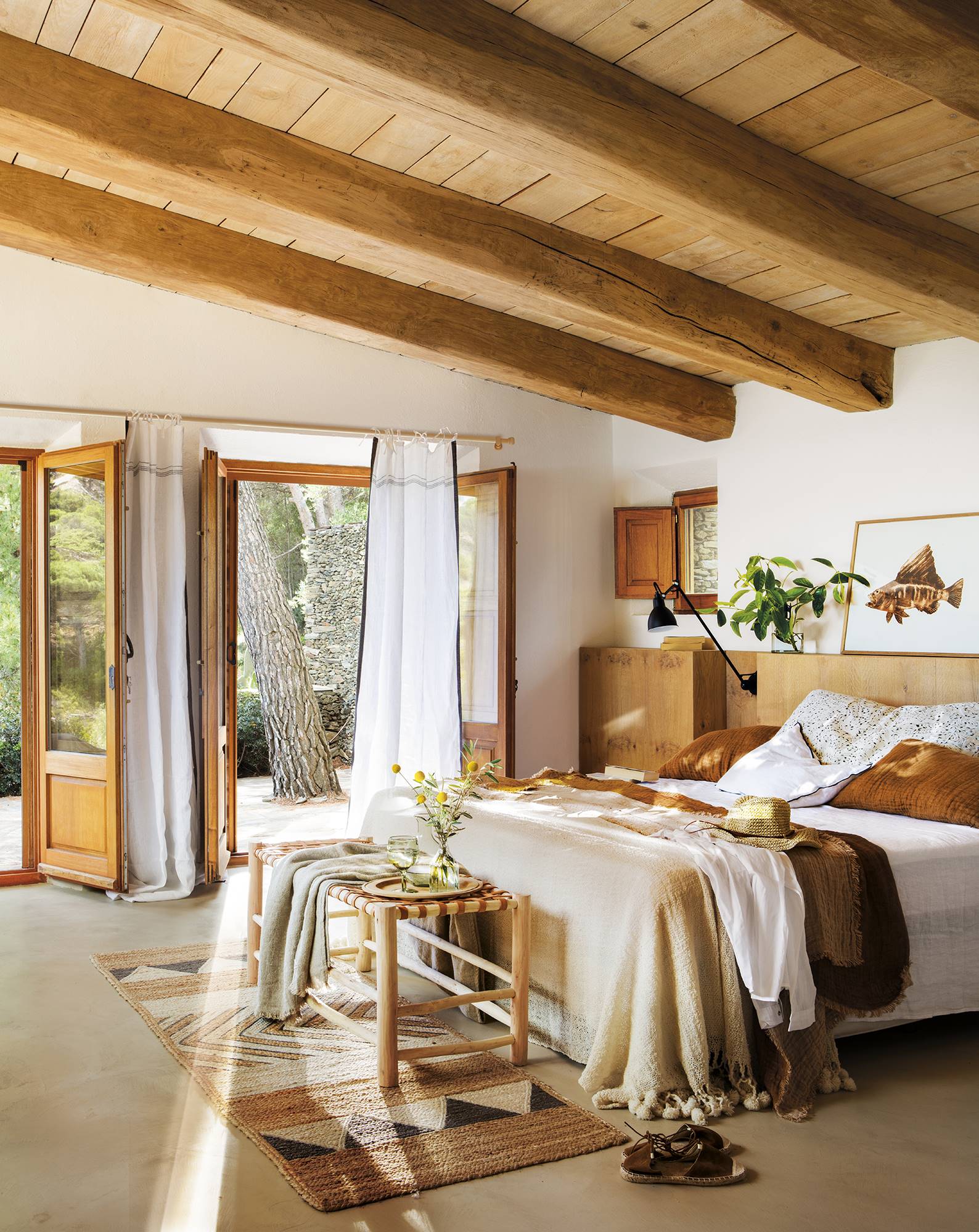 Dormitorio de verano con vigas de madera vistas, cabecero de obra de madera, aplique negro metálico, banco de ratán y alfombra de fibras naturales PANO 1238-1245