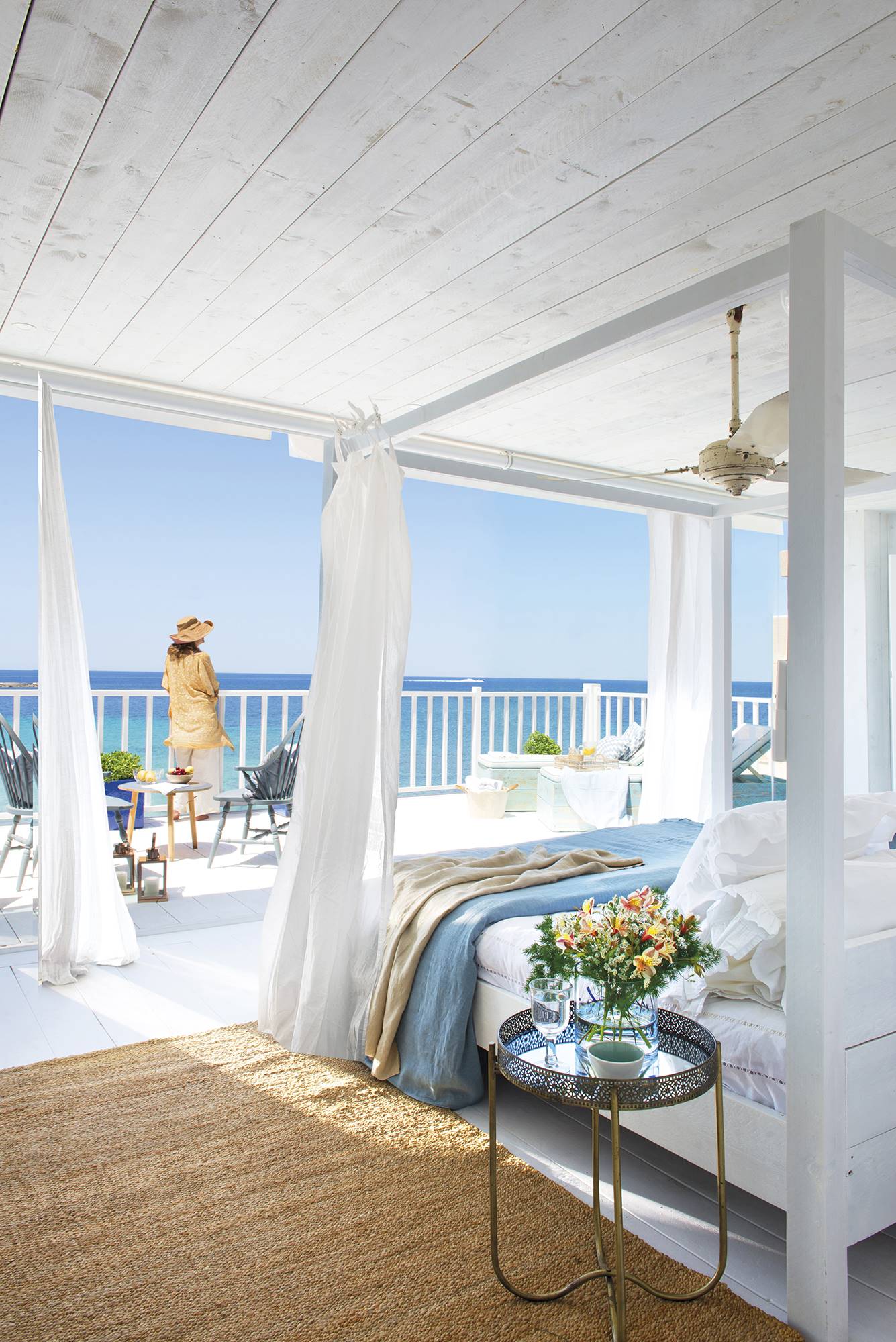 Dormitorio con terraza frente al mar, cama con dosel de madera blanca y ropa de cama en blanco y azul.