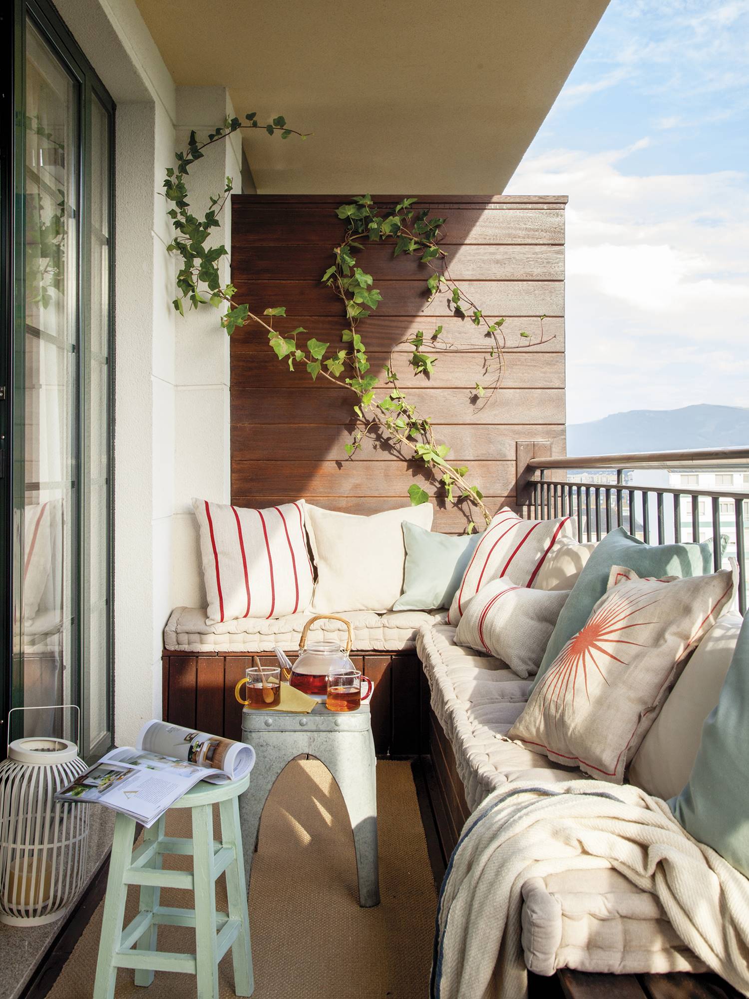 Terraza con banco a medida en L de madera decorado con cojines en tonos beige y azulados y dos mesas auxiliares sobre una alfombra de fibras