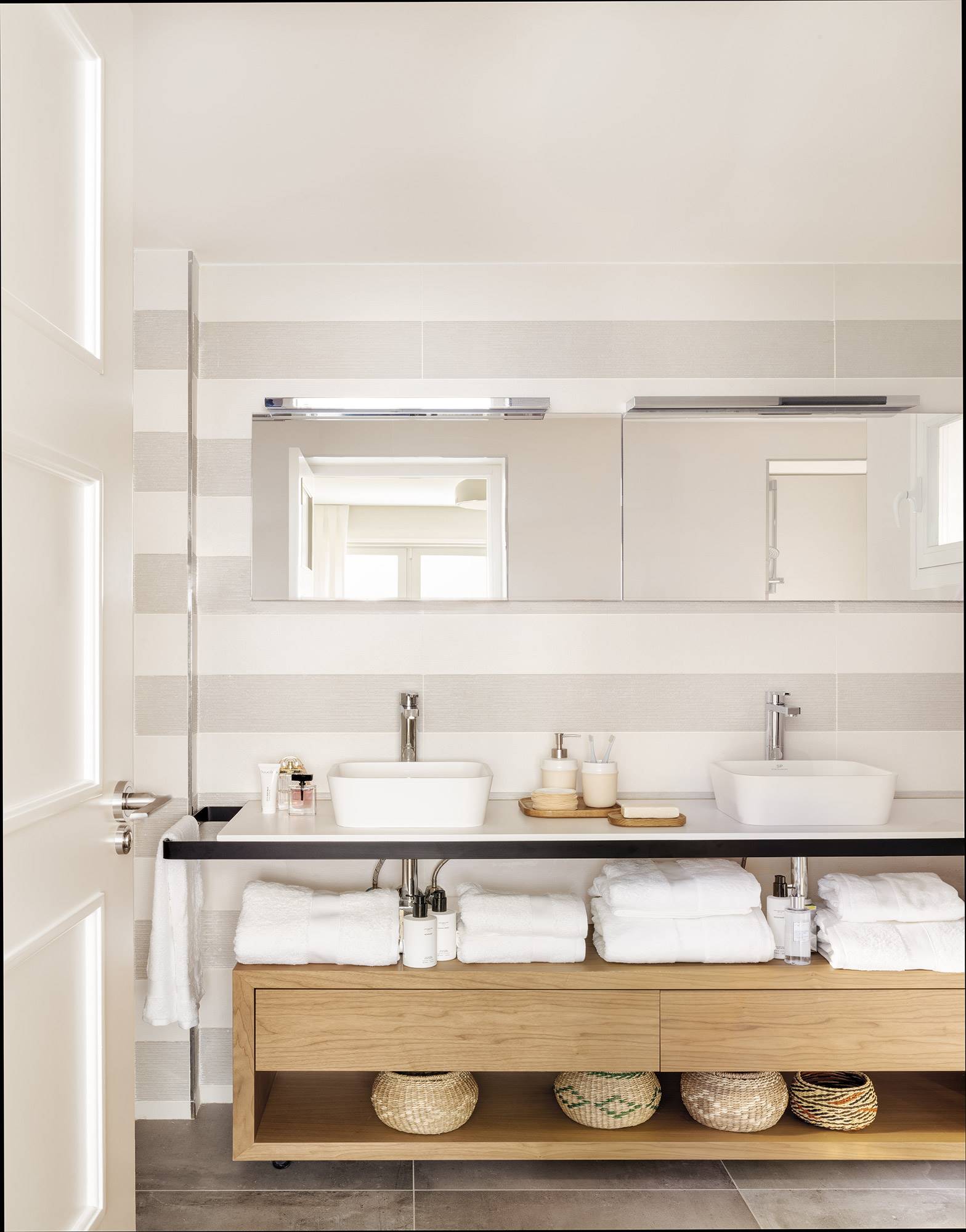 Baño con papel pintado de rayas con lavabos suspendidos y mueble de madera bajo ellos. 