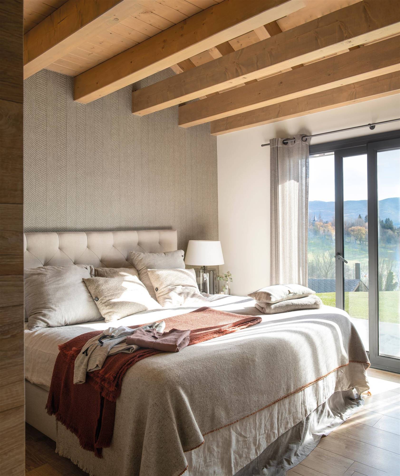 Dormitorio con cabecero tapizado en capitoné, pared revestida de papel pintado gris y vigas de madera.