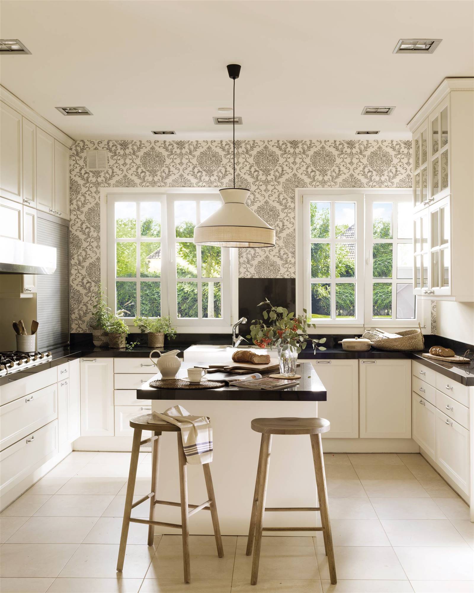 Cocina blanca y negra de estilo clasico con isla y taburetes. Encimera gris antracita y papel pintado en una de las paredes.