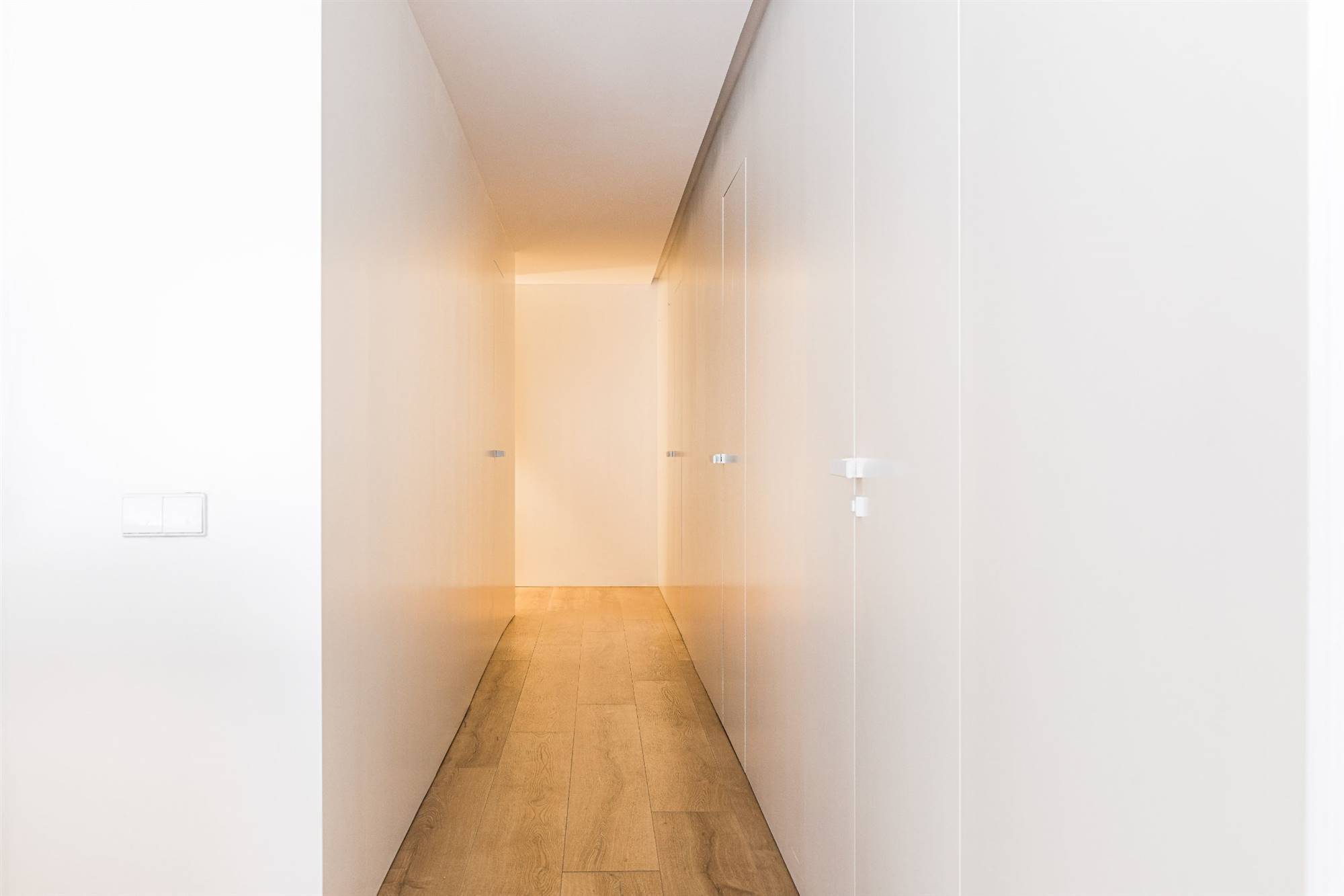 Pasillo moderno y luminoso con suelo de madera, luces leds y paneles lacados en blanco en la pared.