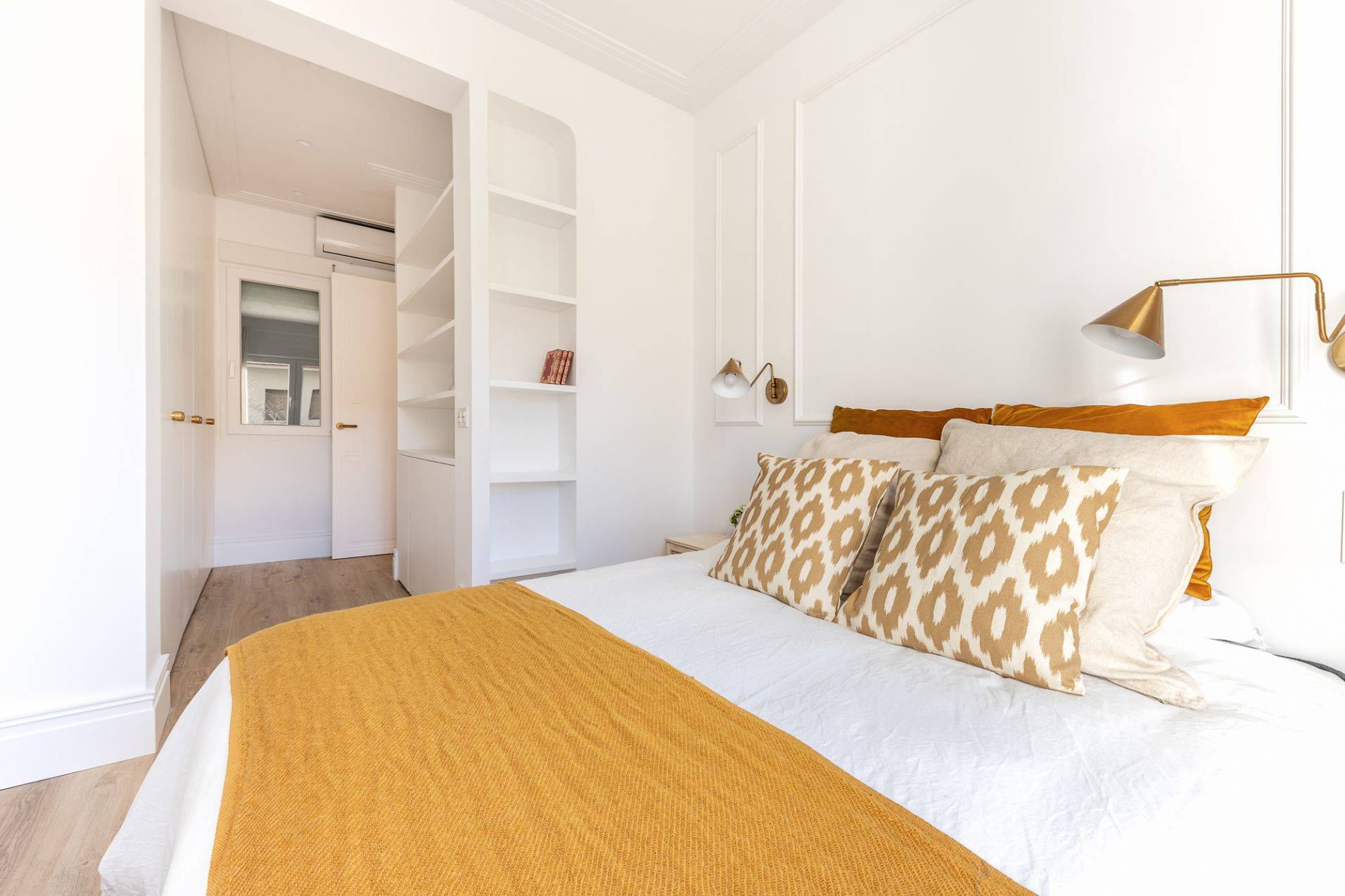 Dormitorio con paredes blancas con molduras, estantería y con ropa de cama mostaza.