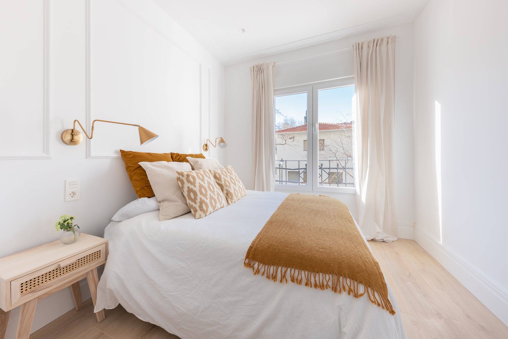 Dormitorio con paredes blancas con molduras, ventana con cortina, mesita de noche de madera y ropa de cama en tonos tierra. 