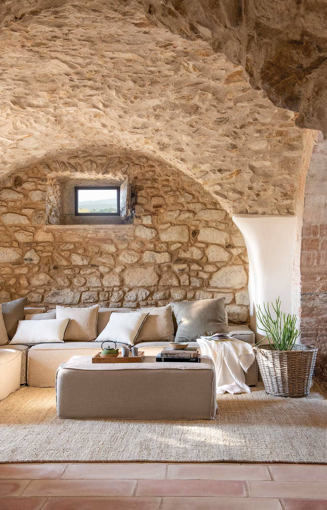 Salón rústico y minimalista con paredes de piedra, sofá beige a juego con el reposapiés y alfombra de fibras 00538550