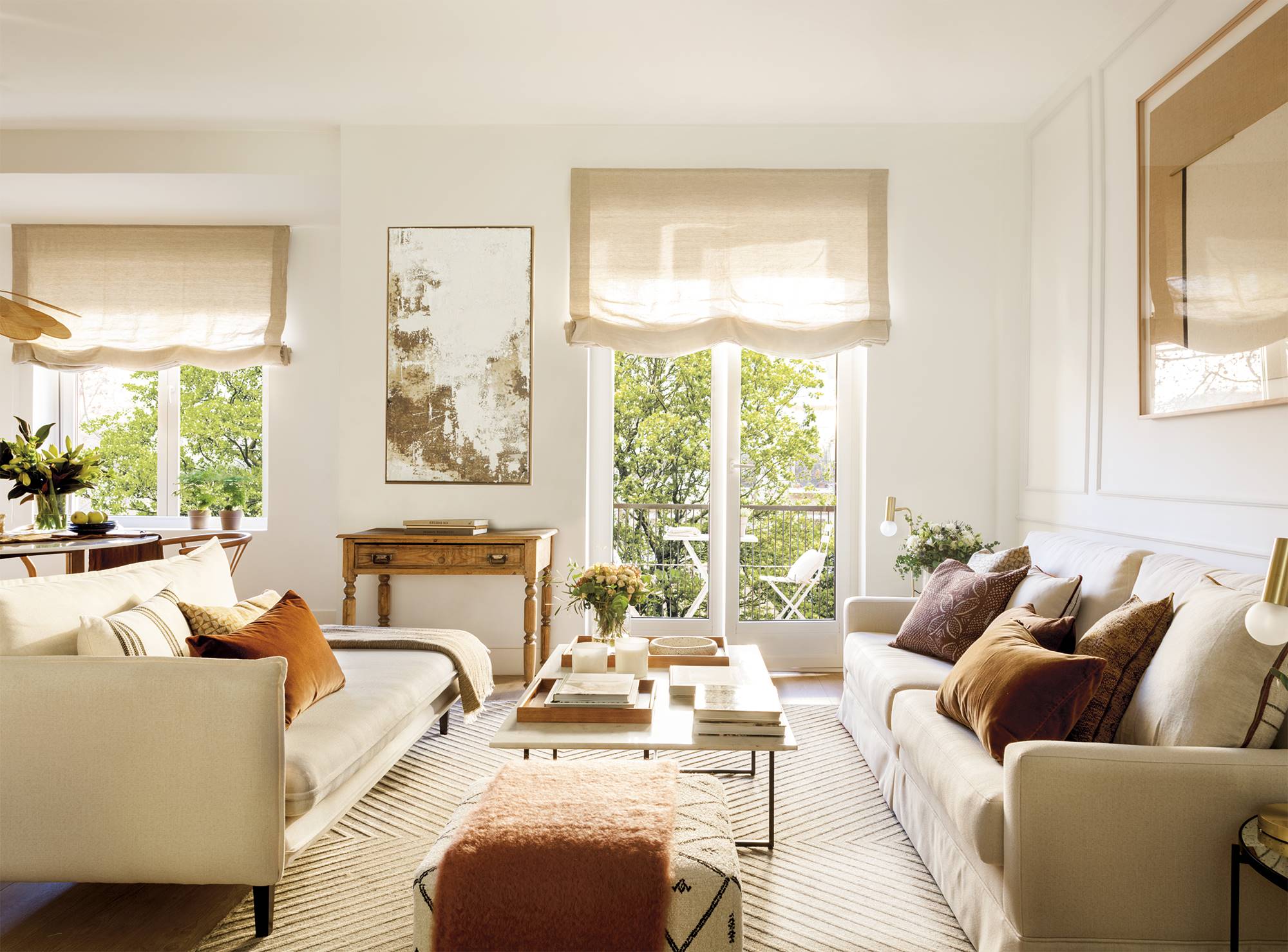 Sala de estar moderna con sofá, puf y estores en color crema, y cojines y cuadros en colores tierra. 