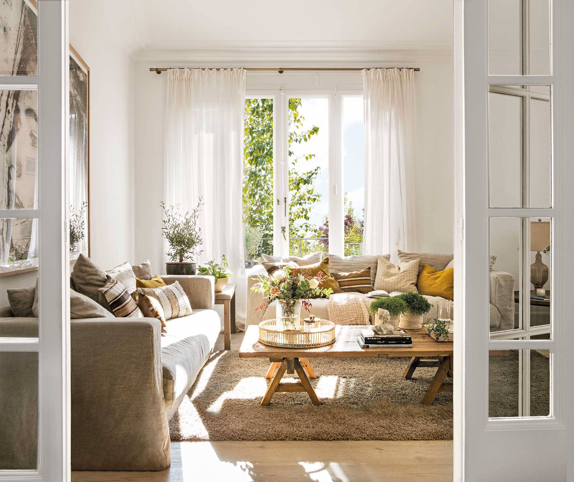 Salón blanco con mesa de centro de madera, alfombra de pelo, dos sofás y ventana con cortinas.