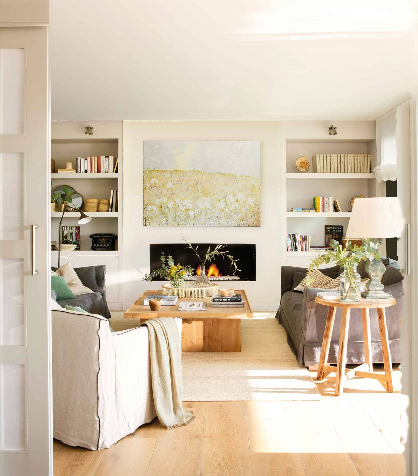 Salón con chimenea en tonos blanco roto, sofá gris, cuadro grande y estanterías a ambos lados.
