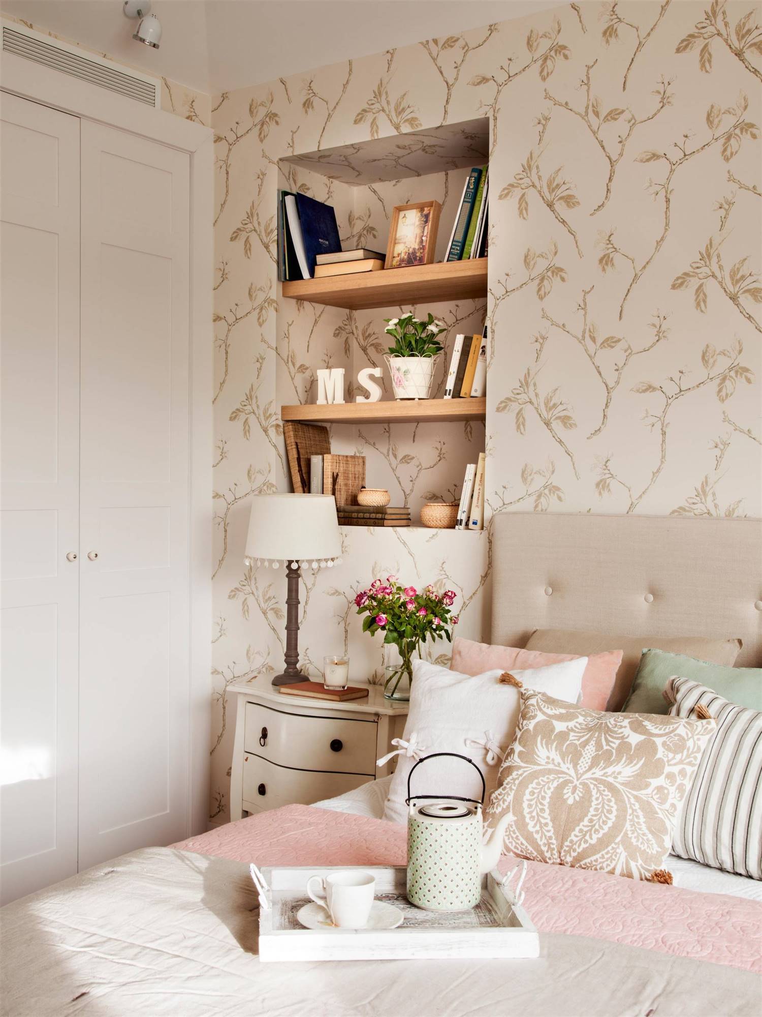 Dormitorio con cabecero de capitoné, cojines, estantería y papel pintado. 
