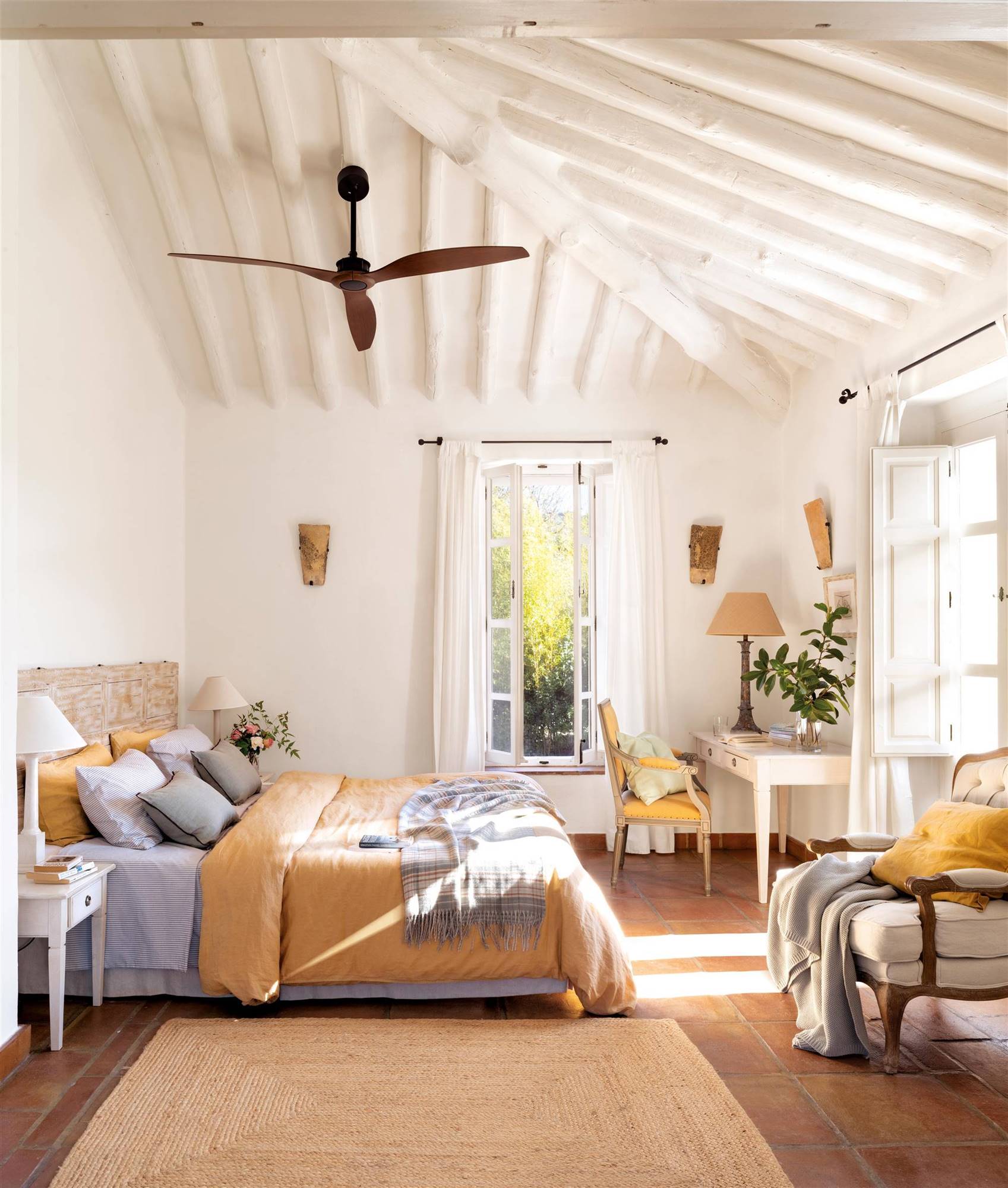 Dormitorio de verano con techos abuhardillados blancos, ventilador de techo y textiles en color mostaza. 
