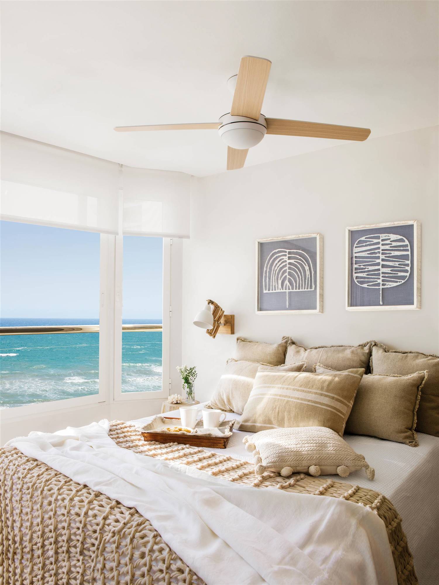 Dormitorio de verano con vistas al mar, ventilador de techo y ropa de cama blanca y beige. 