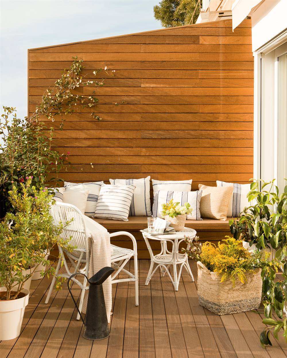 Pequeña terraza con estar, pared con lamas de madera, suelo entarimado, banco, cojines a rayas y sillas de fibras vegetales