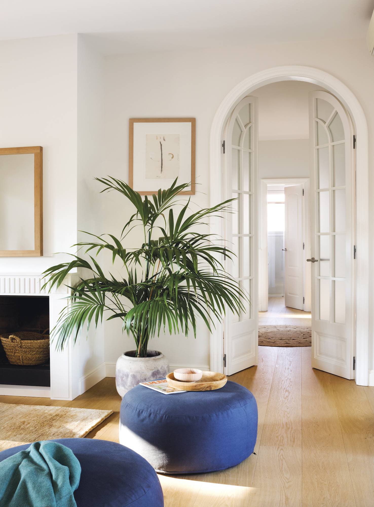 Rincón de un salón blanco con una palmera grande, pufs azules, chimenea y puerta de cristal. 