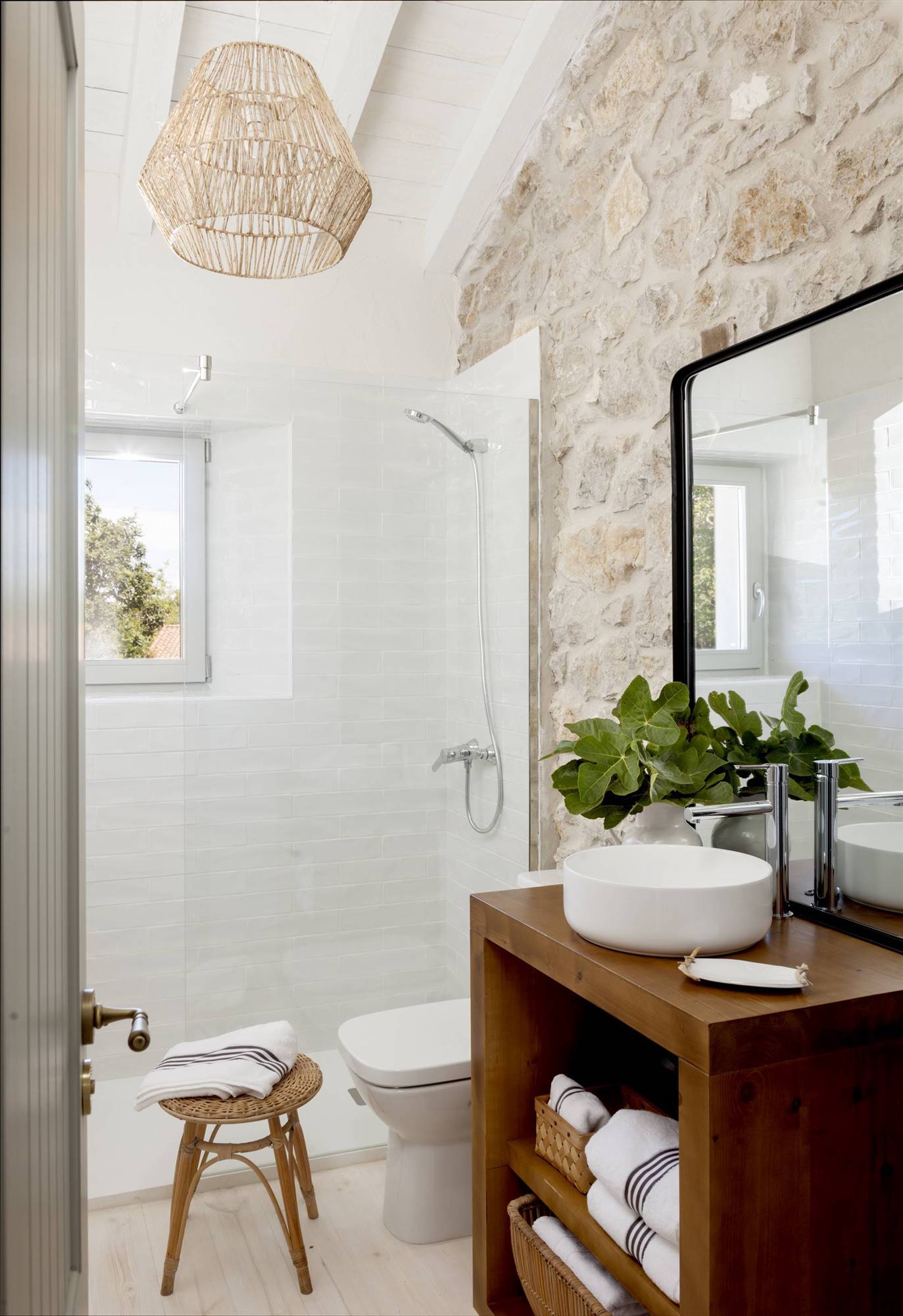 Baño rústico con pared de piedra, azulejos blancos en la ducha y mueble de madera. 
