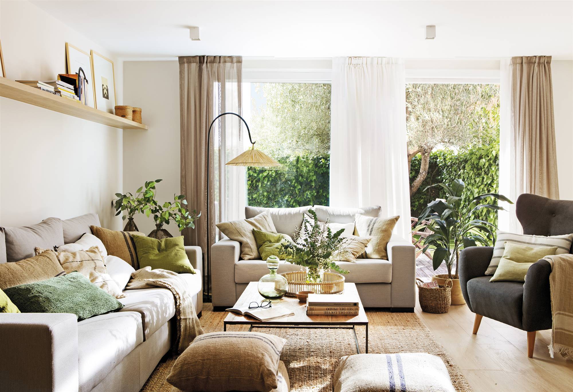 Salón decorado con plantas, con dos sofás grises, butaca orejera, puffs, lámpara de pie y alfombra de yute. 