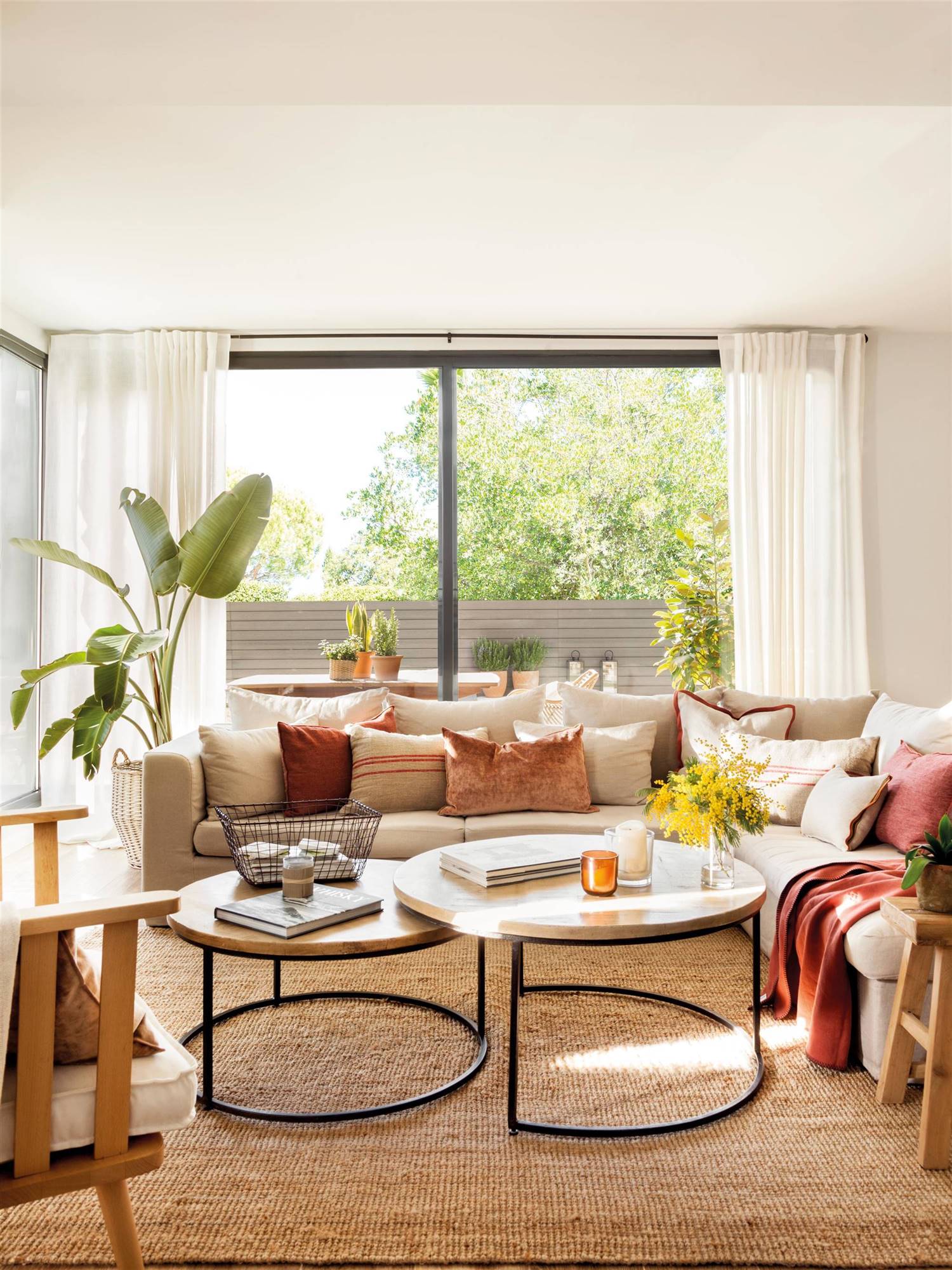 Salón con sofá en forma de L, mesas redondas tipo nido, terraza y plantas.